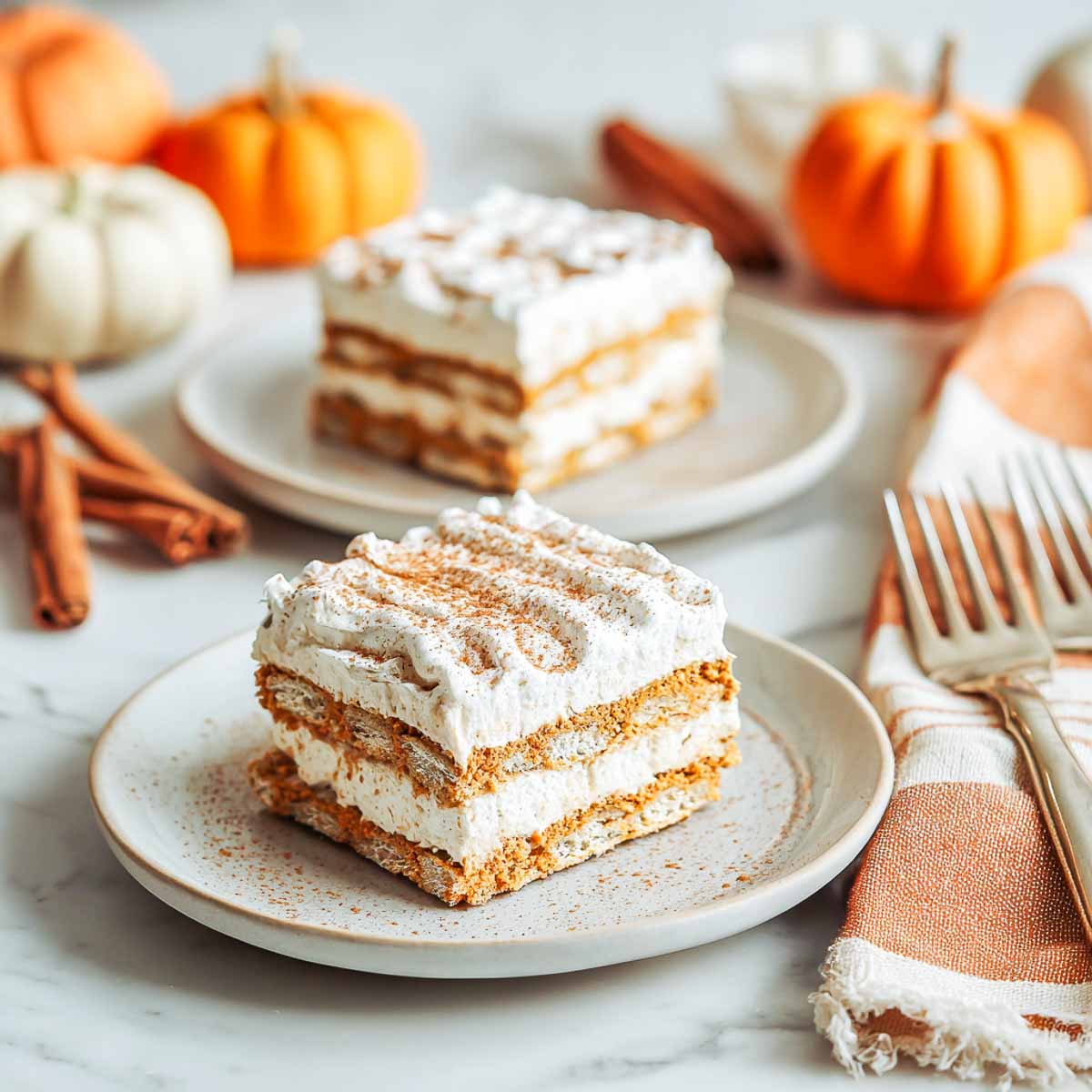 Two plates with square slices of gluten-free pumpkin pie icebox cake, layered with cookies, pumpkin filling, and whipped topping, sprinkled with cinnamon.