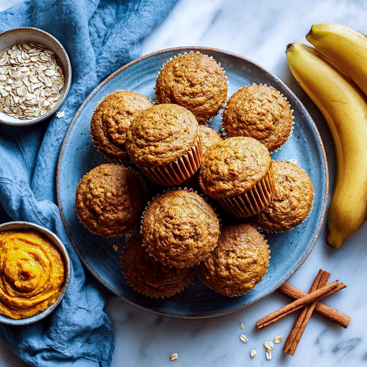 Pumpkin banana oatmeal muffins on ceramic plate with bananas, pumpkin puree, oats, and cinnamon sticks.