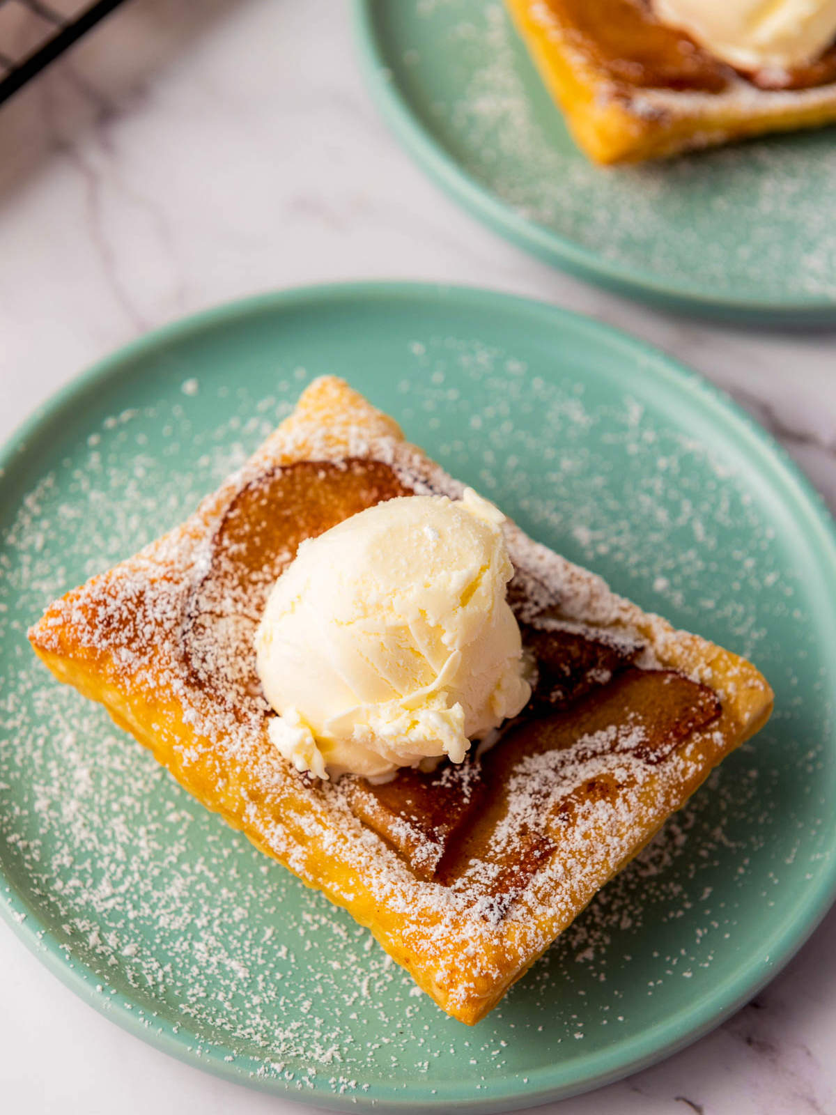 Close-up of an upside down apple tart with caramelized apples, a scoop of vanilla ice cream, and powdered sugar on a teal plate.