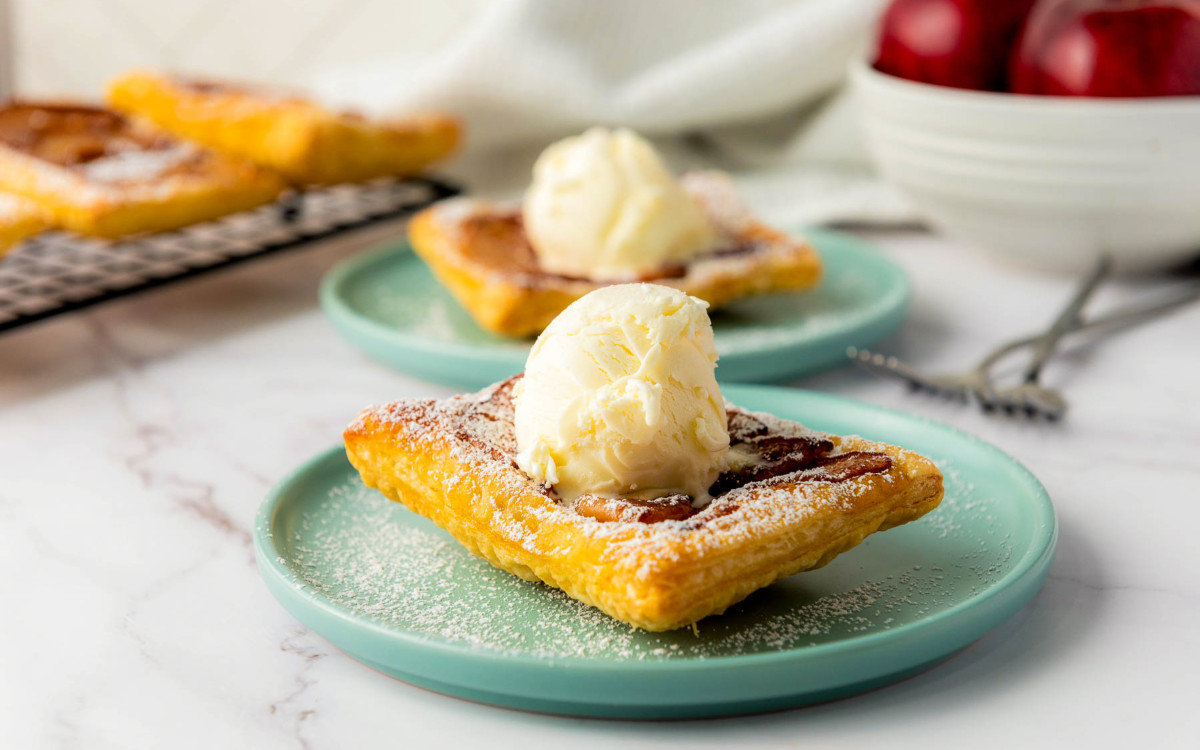 Side view of upside down apple tart with golden puff pastry, vanilla ice cream scoop, and powdered sugar on a teal plate.