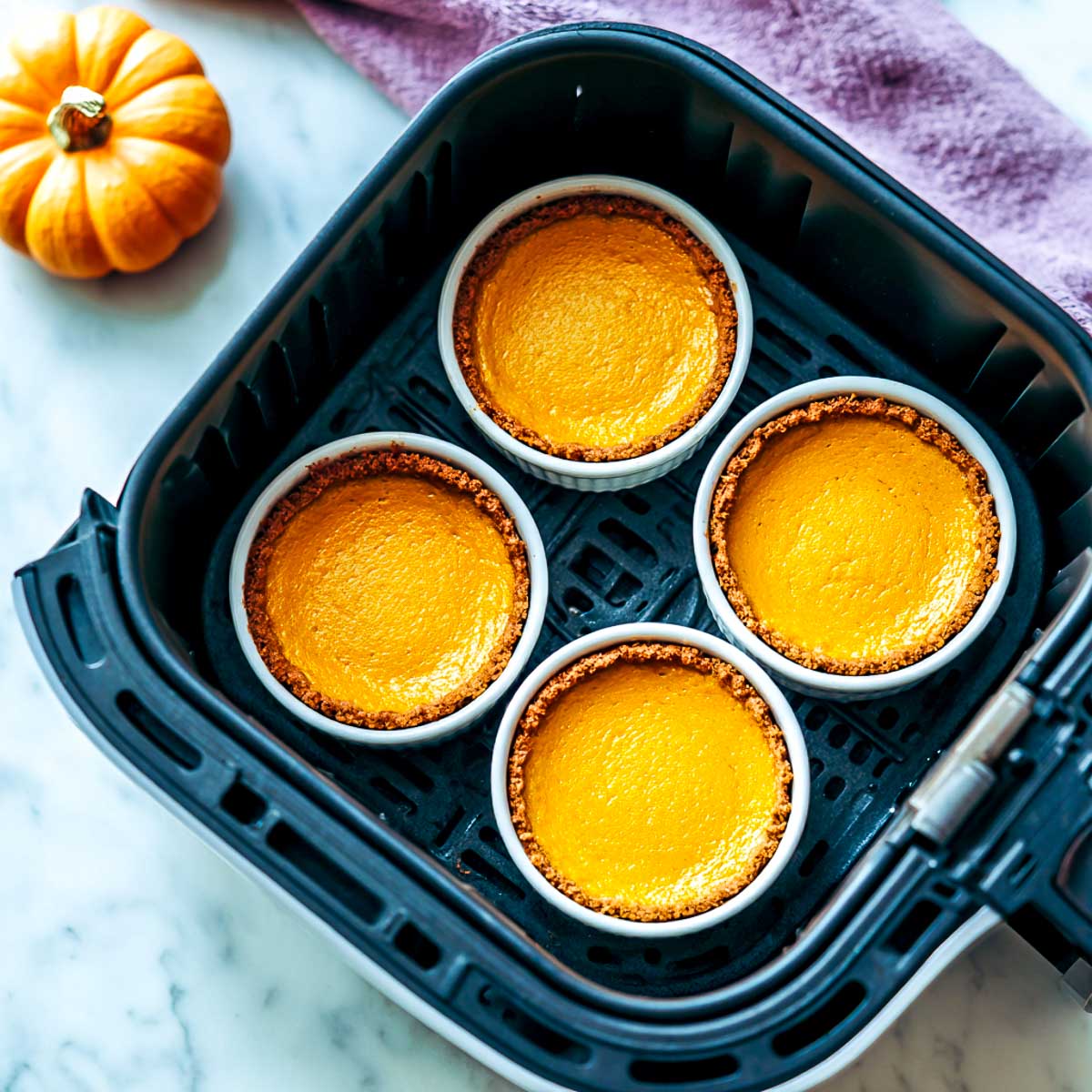 Four mini pumpkin pies baking inside air fryer basket with purple napkin.