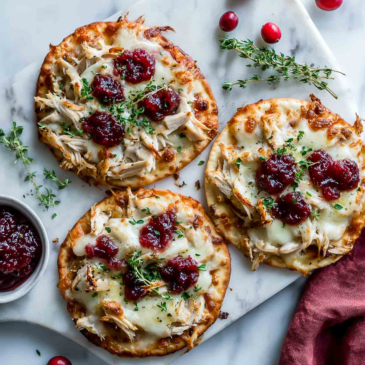 Angled view of mini turkey and cranberry flatbread pizzas topped with melted mozzarella, cranberry sauce, and fresh thyme.