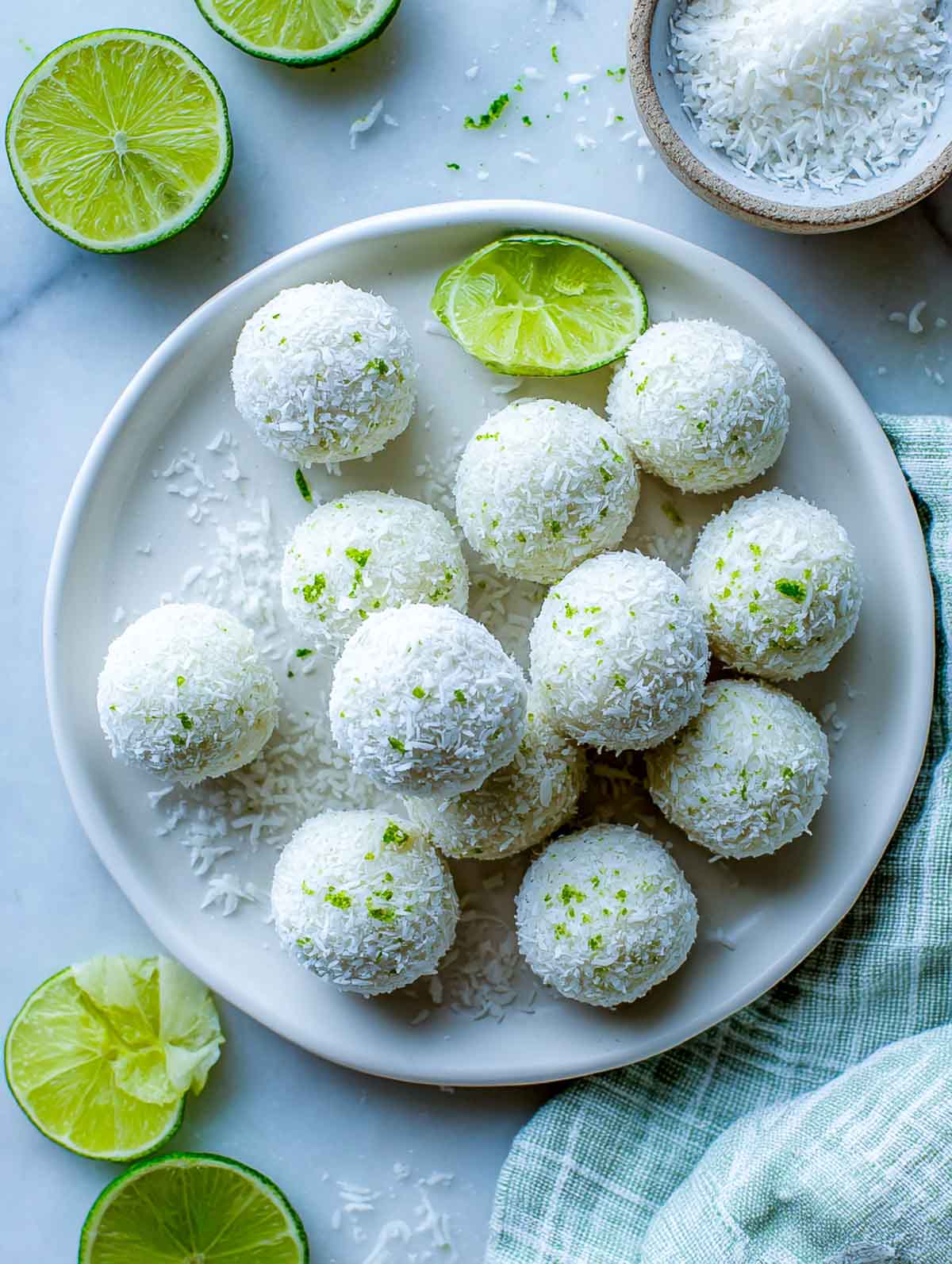 Coconut lime energy bites on a white plate with lime halves, zest, and shredded coconut on a marble surface.