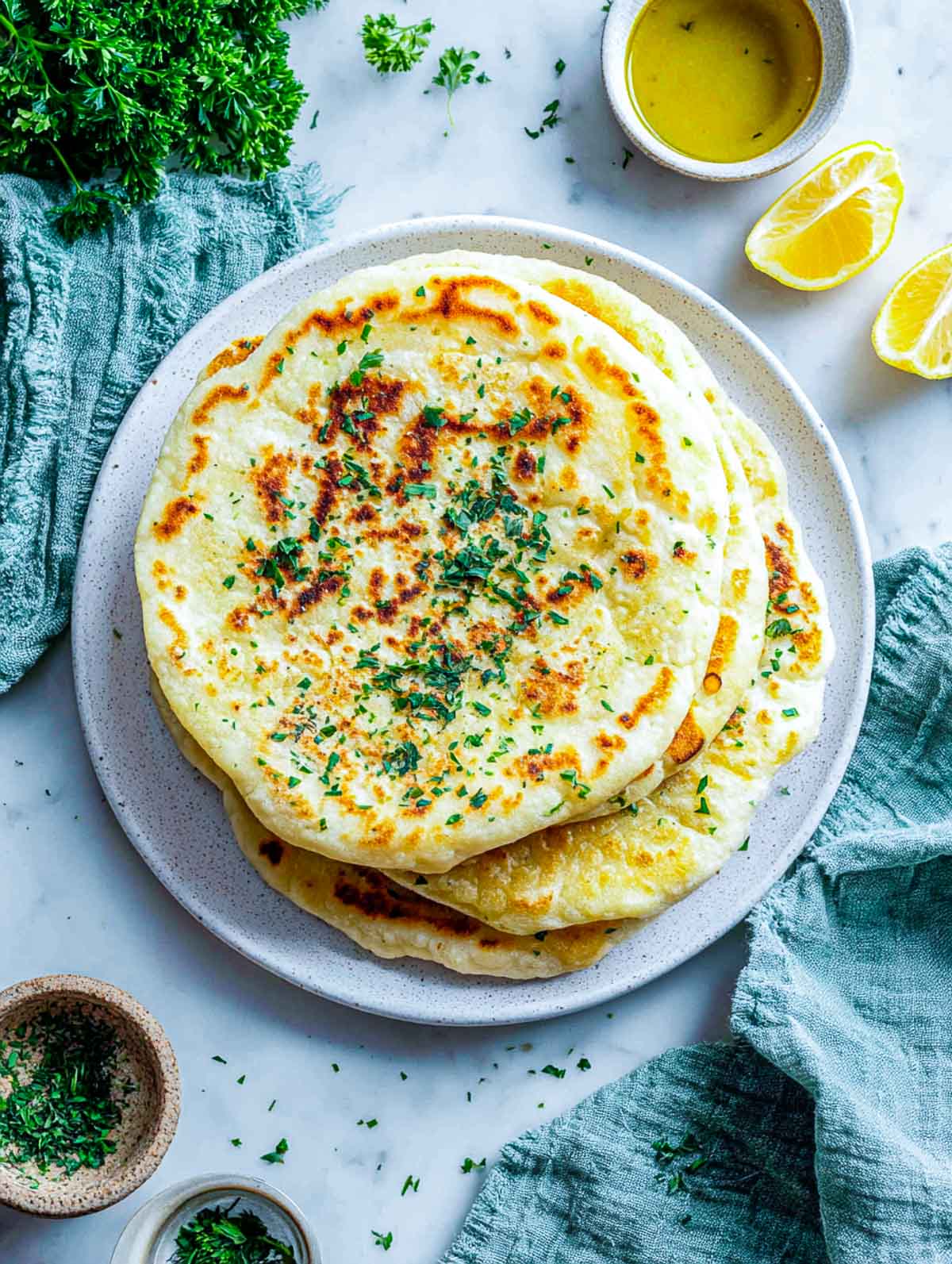 Golden 3-Ingredient Greek Yogurt Flatbreads stacked on a white plate with herbs, lemon wedges, and olive oil on a marble background.