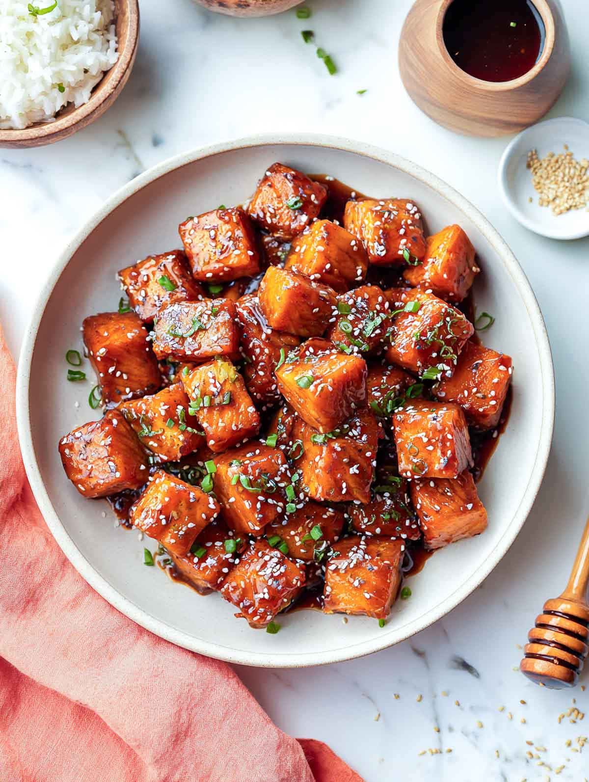 Golden honey soy salmon bites sprinkled with sesame seeds and scallions in a shallow bowl with rice on the side.