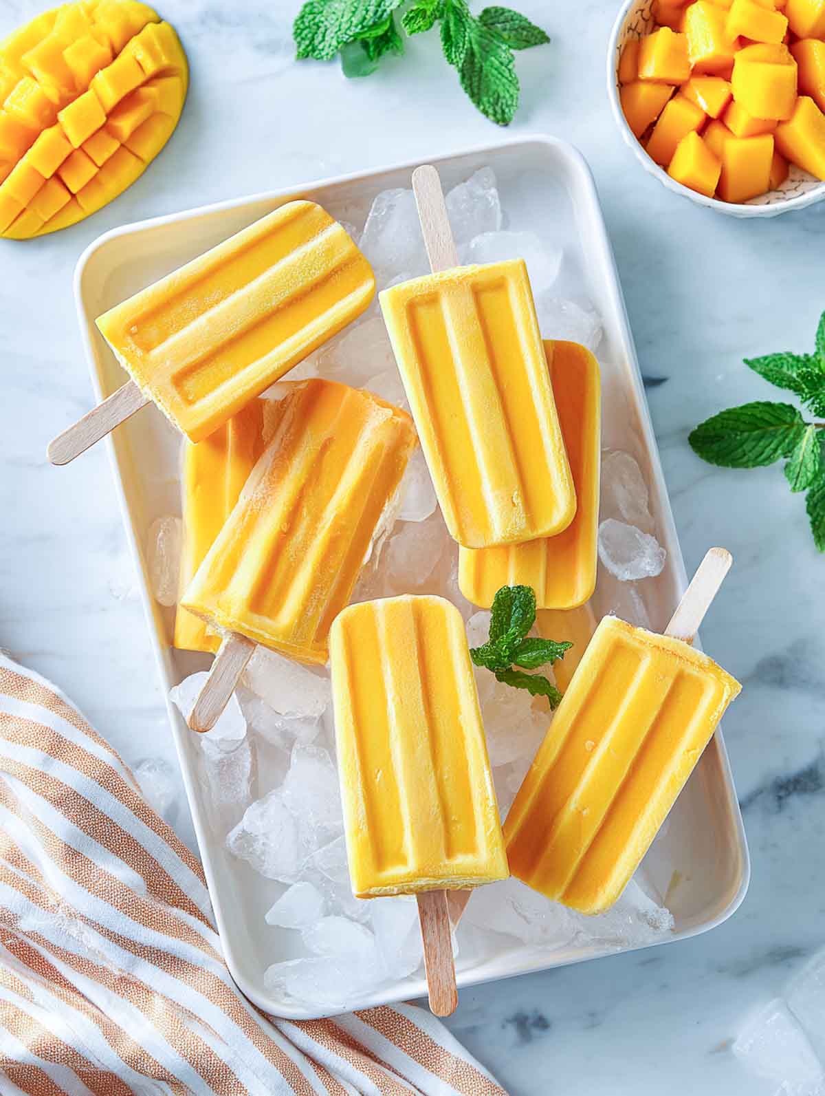 Creamy mango popsicles on a bed of ice with mint and diced mango on a marble counter.