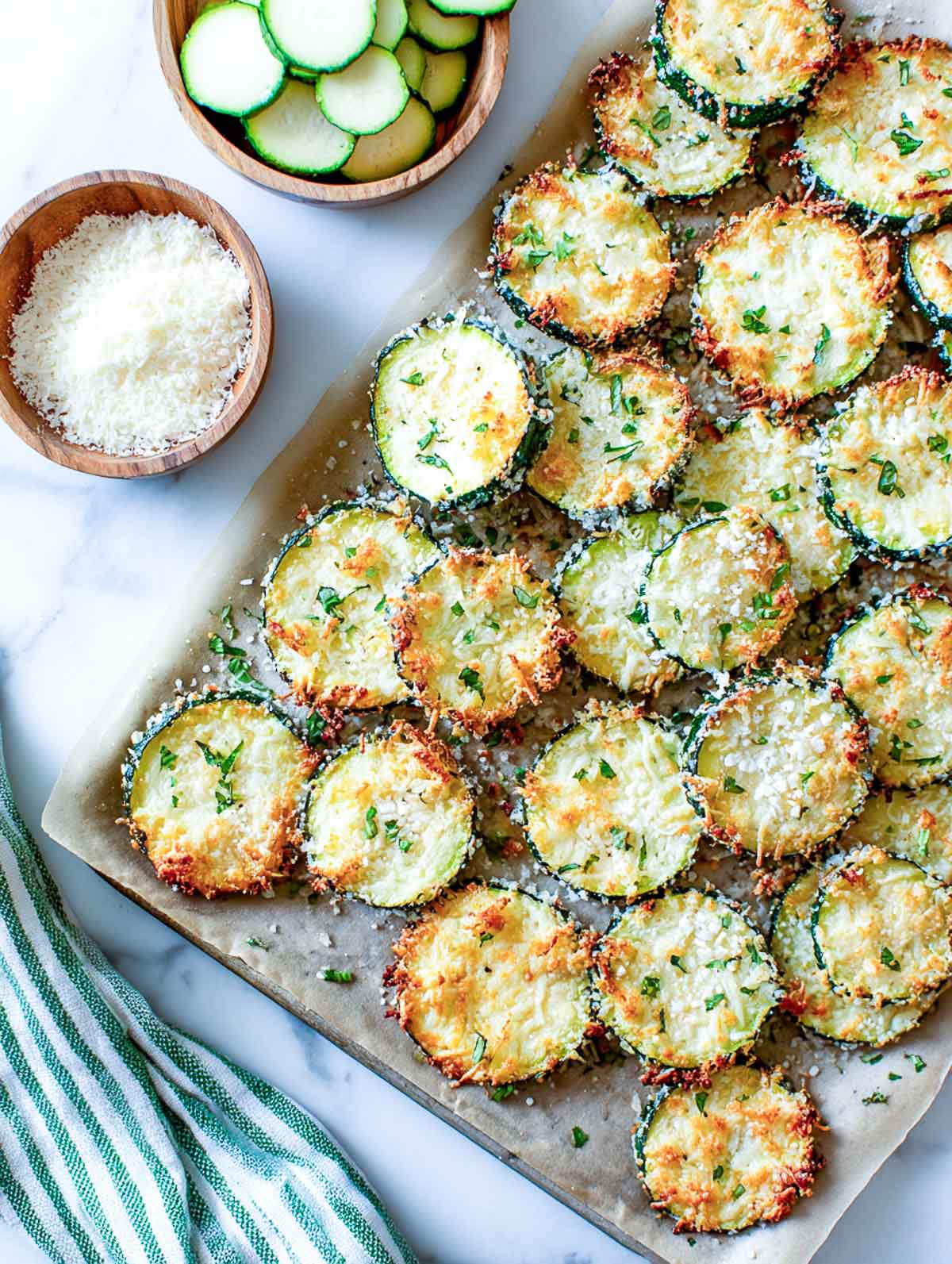 Golden parmesan zucchini chips on a parchment-lined tray with bowls of grated cheese and sliced zucchini beside a green-striped napkin.