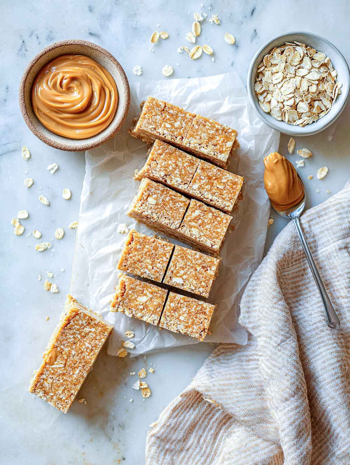 Peanut butter oat bars stacked on parchment paper with a bowl of oats and creamy peanut butter beside them.