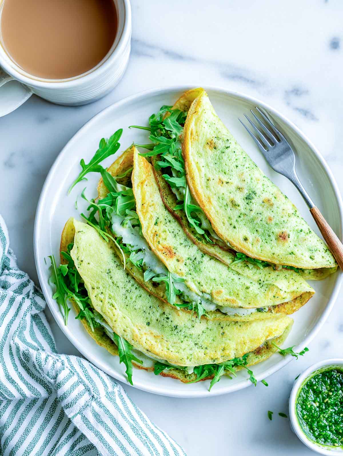 Pesto egg wraps folded on a white plate with arugula and melted cheese, coffee mug and green napkin on a marble counter.