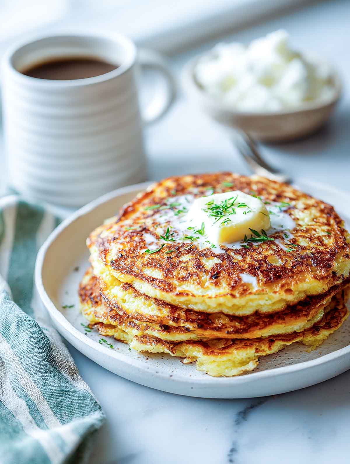 Stack of savory cottage cheese pancakes on a white plate topped with butter and herbs, with coffee and a napkin beside.