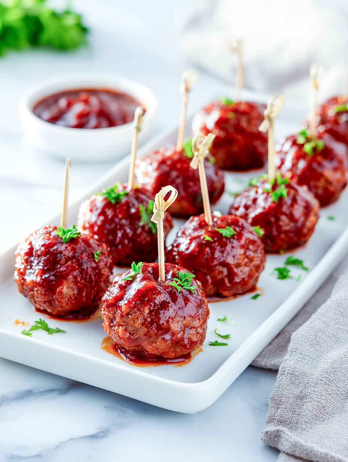 BBQ meatballs arranged on a rectangular white plate with food picks, garnished with parsley and served with a small bowl of BBQ sauce.