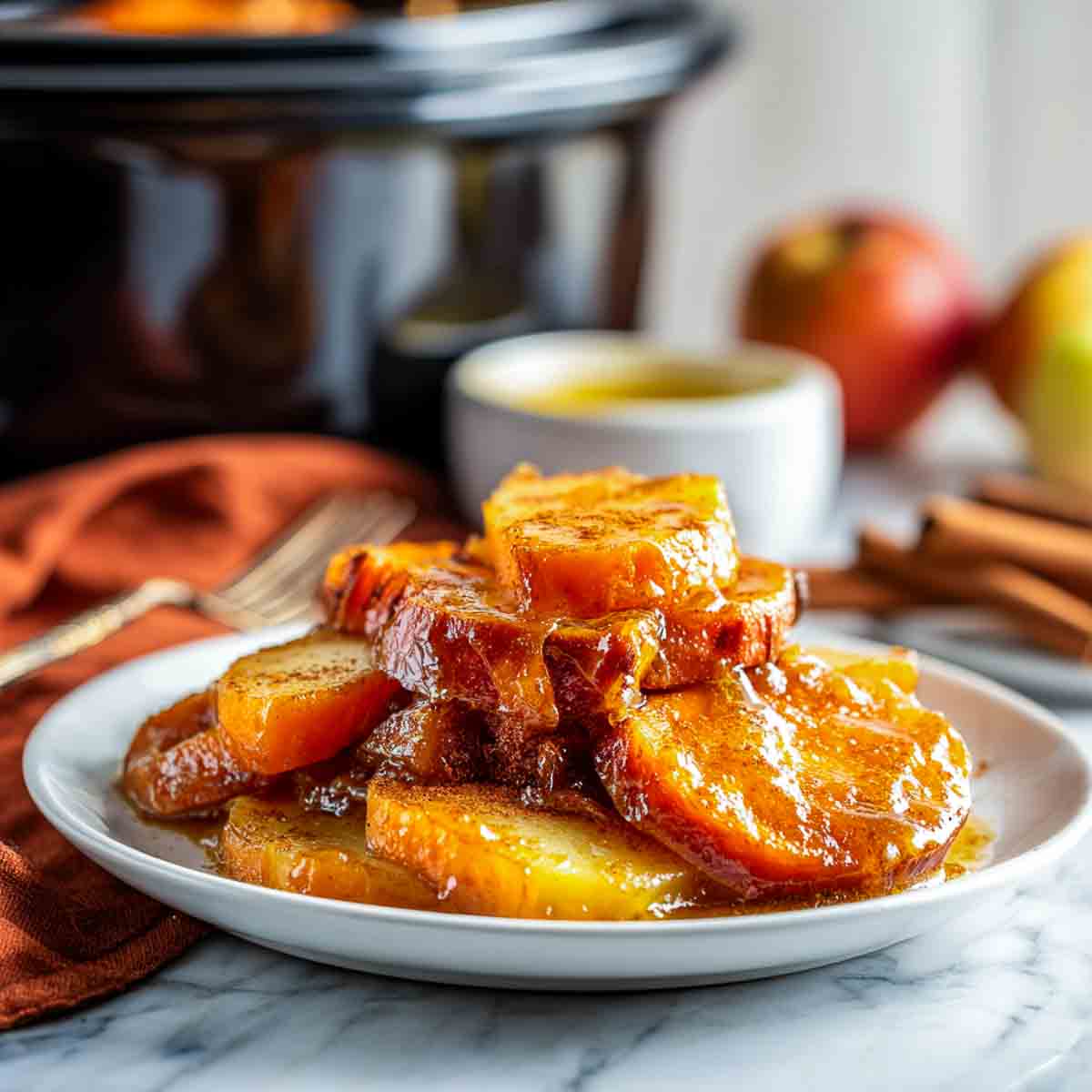 A plated serving of crockpot sweet potato and apple casserole drizzled with cinnamon butter glaze, with apples, cinnamon sticks, and a black crockpot in the background.
