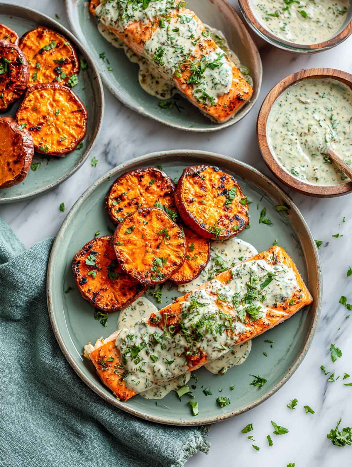 Top-down view of two blue ceramic plates with grilled sweet potato rounds and salmon fillets topped with creamy herbed yogurt sauce.