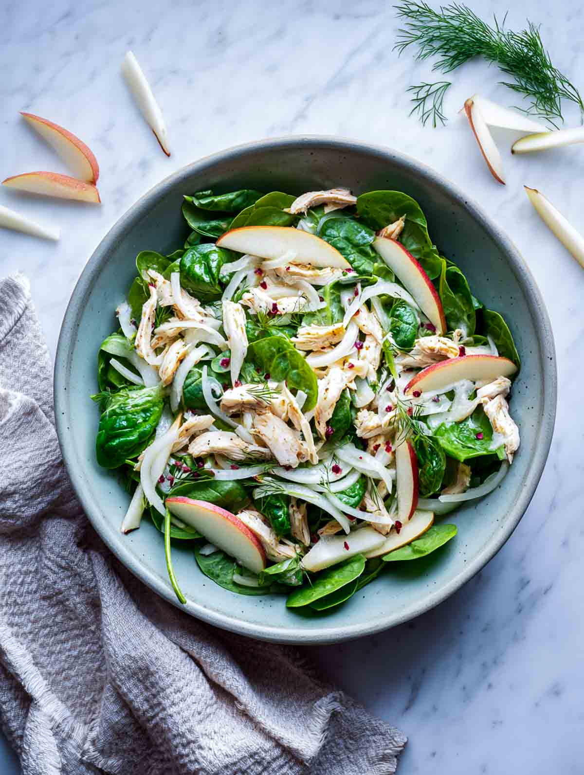 Spinach chicken salad with fennel, pear slices, shredded chicken, and cranberry dressing in a serving bowl placed on a marble surface.