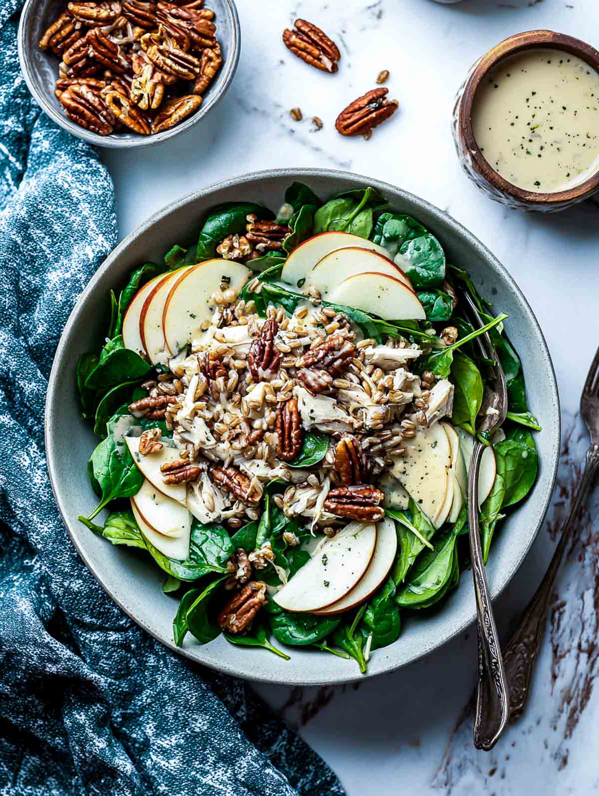 Top-down view of a winter power salad in a muted stoneware bowl filled with shredded chicken, fresh spinach, thin apple slices, toasted pecans, and farro, topped with apple cider dressing.