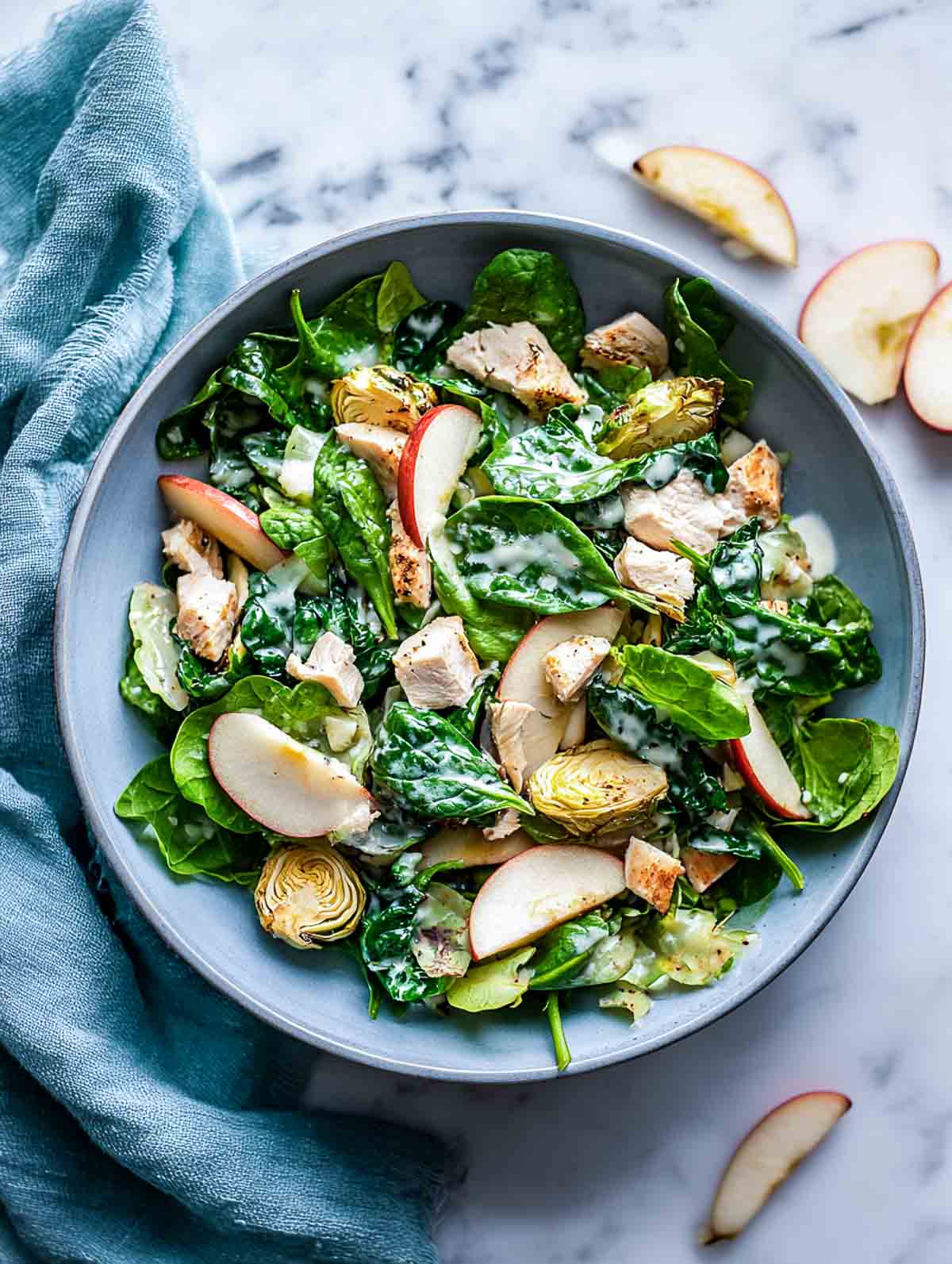 A top-down view of a winter salad with chopped chicken, roasted Brussels sprouts, spinach, and apple chips arranged in a muted grey-blue bowl on a marble surface.