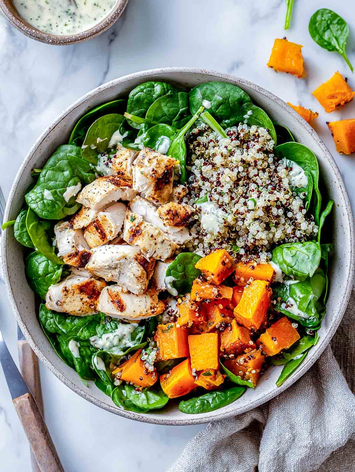 A top-down view of a winter salad with roasted butternut squash cubes, quinoa, spinach, and rustic slices of chicken arranged in a muted ceramic bowl on a marble surface.