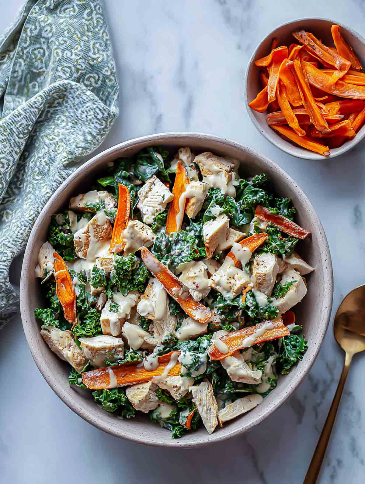 A top-down view of a spinach chicken salad topped with roasted carrot ribbons and a creamy lemon-tahini drizzle, served in a muted beige bowl.