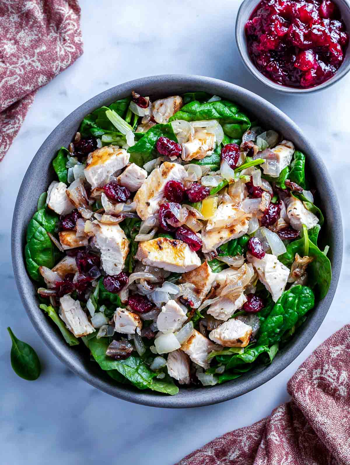 A top-down view of a spinach chicken salad topped with chopped chicken, roasted shallots, and spoonfuls of cranberry-apple relish.