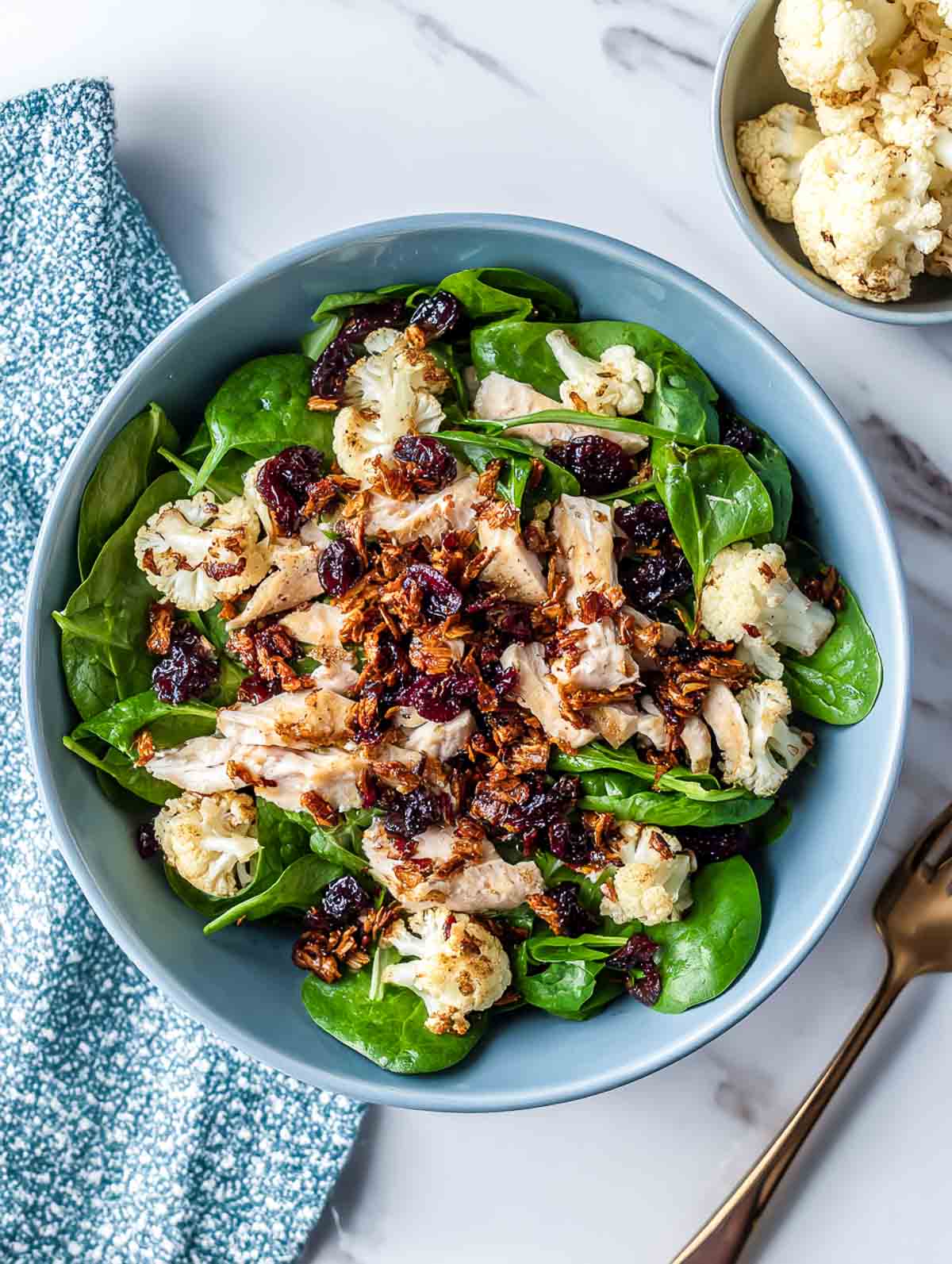 A top-down view of a spinach chicken salad topped with chopped chicken, roasted cauliflower florets, dried cranberries, and a mild chili crunch.