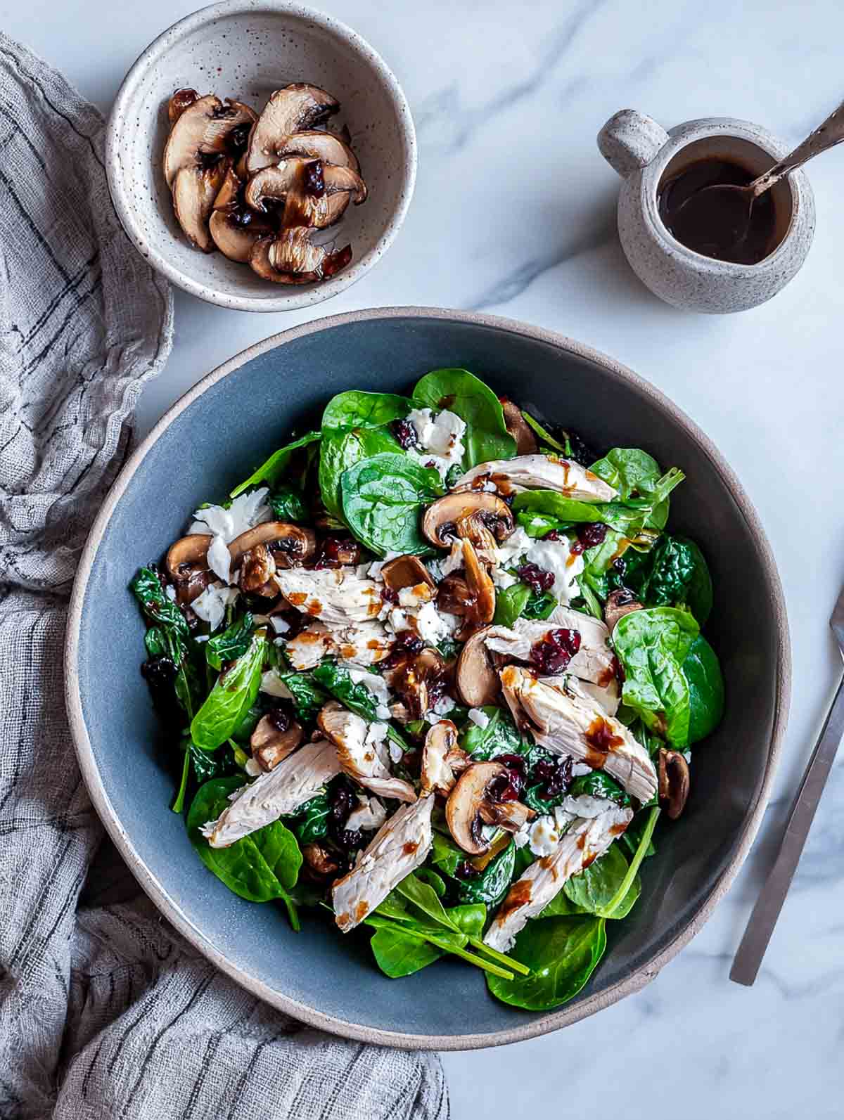 A top-down view of a winter salad with torn chicken pieces, roasted mushrooms, spinach, and a balsamic-cranberry glaze arranged in a muted slate bowl on a marble surface with a small bowl of roasted mushrooms.