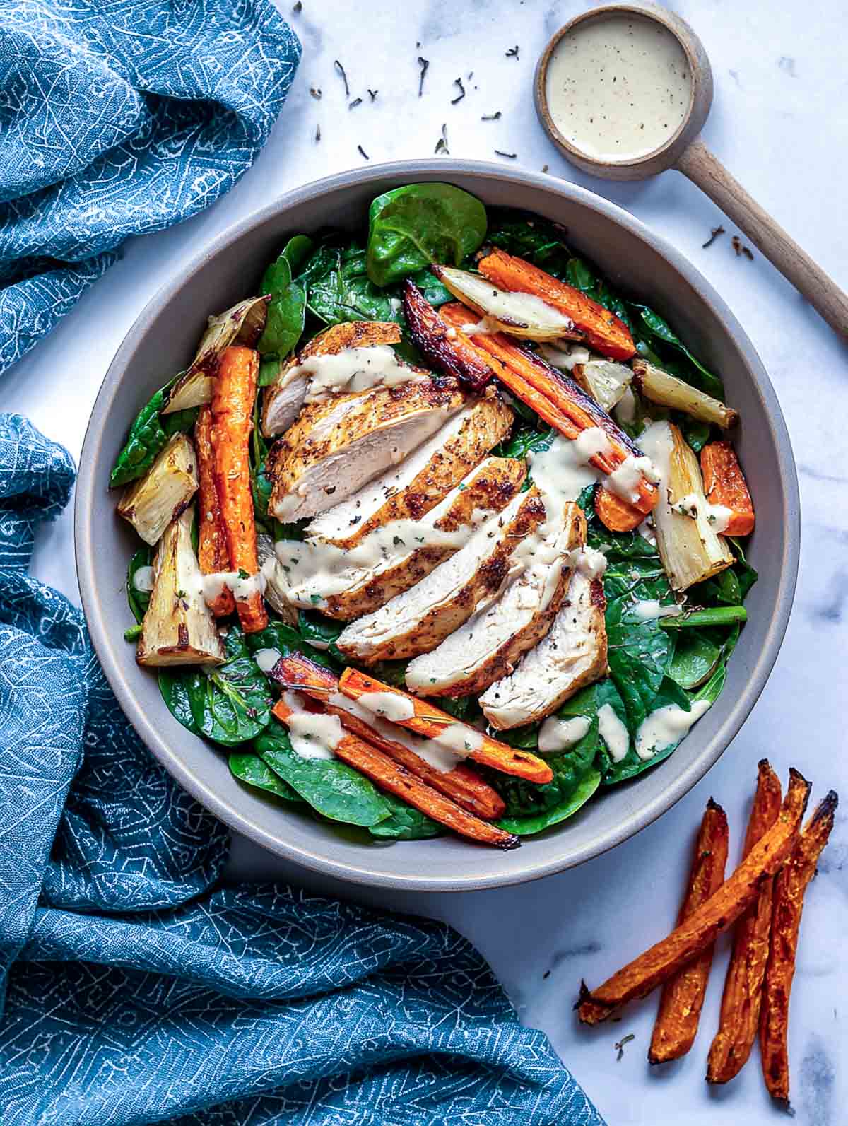 Top-down view of a spinach chicken salad in a neutral stoneware bowl filled with sliced roasted garlic chicken breast, roasted carrots and parsnips, fresh spinach, and a light drizzle of creamy dressing.