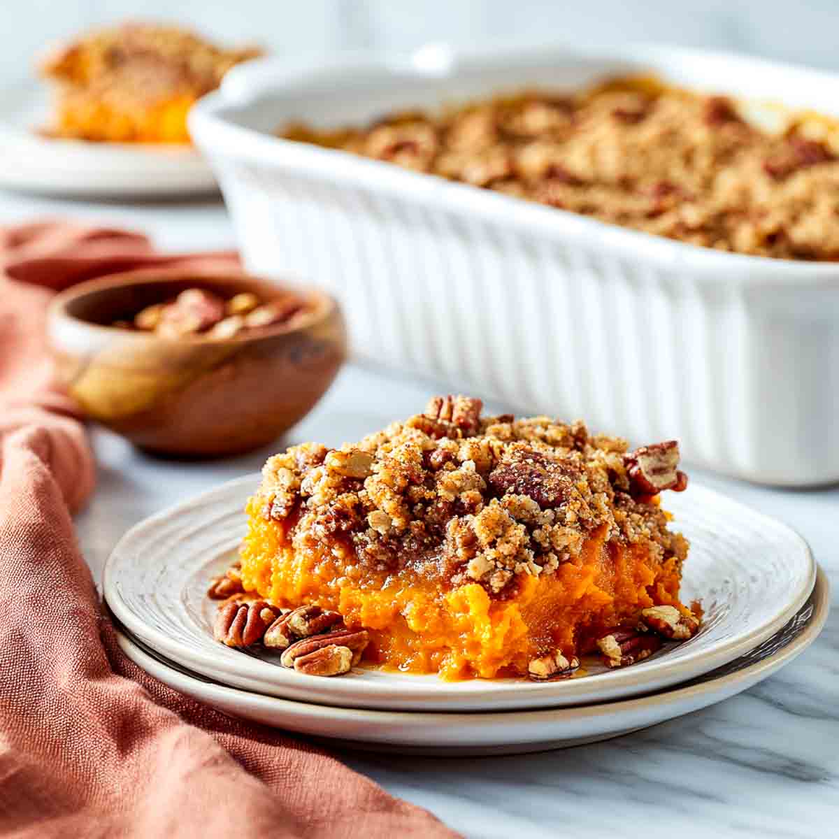 A plated serving of sweet potato casserole topped with oat pecan streusel, with the baking dish in the background on a marble surface.