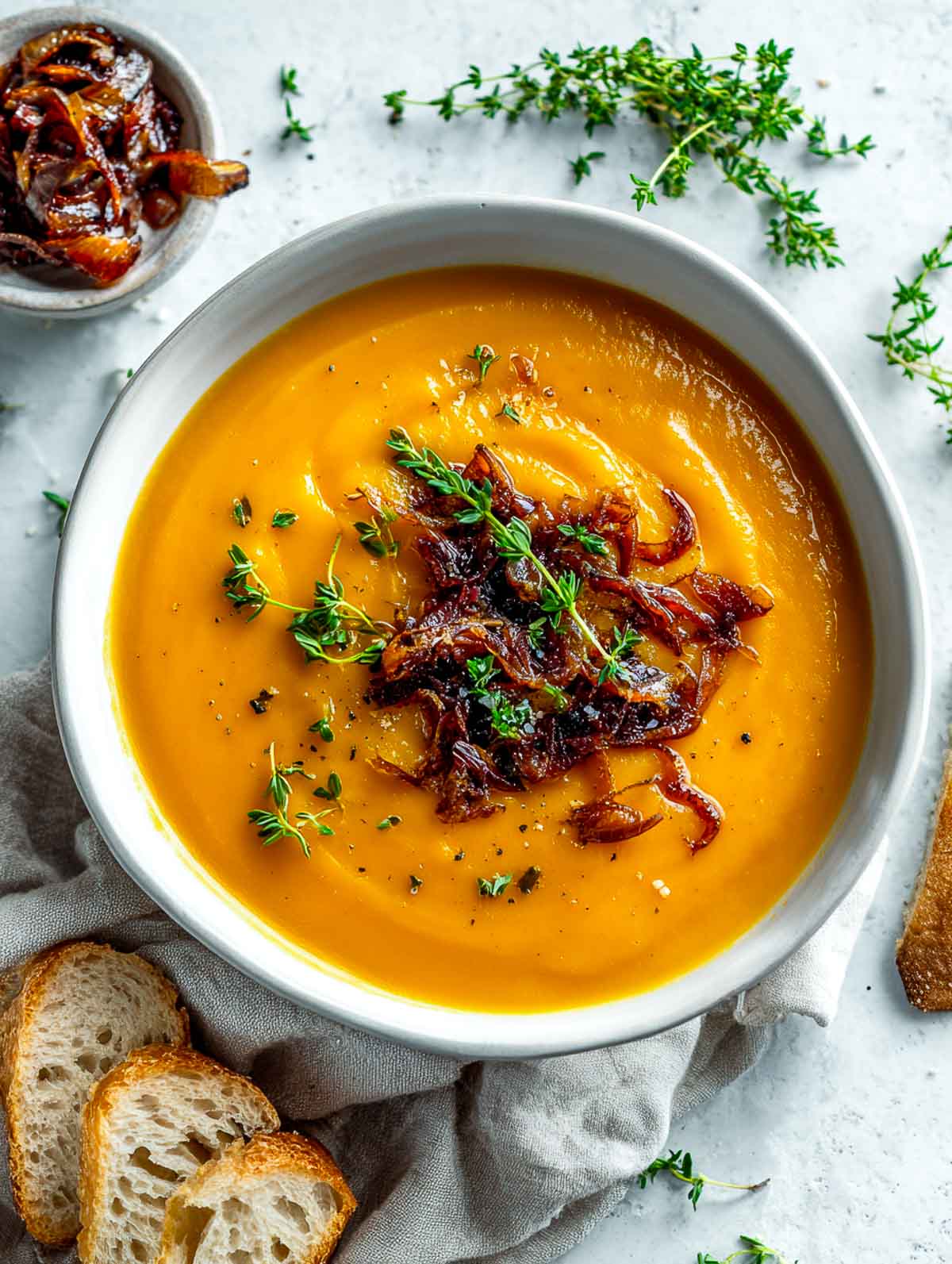 Creamy sweet potato soup topped with caramelized onions and thyme, served with crusty bread on a marble surface.