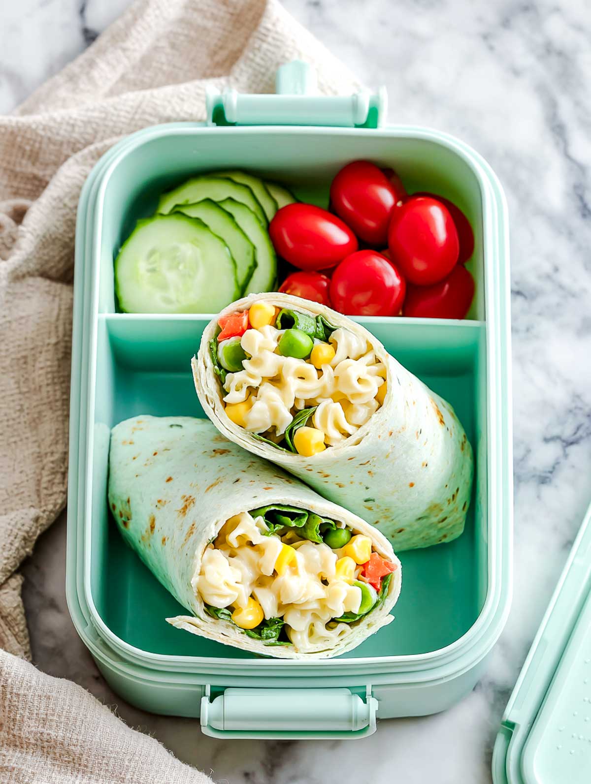 Veggie-packed cold pasta wraps in a mint-green lunchbox with cucumber slices and cherry tomatoes on a marble surface.