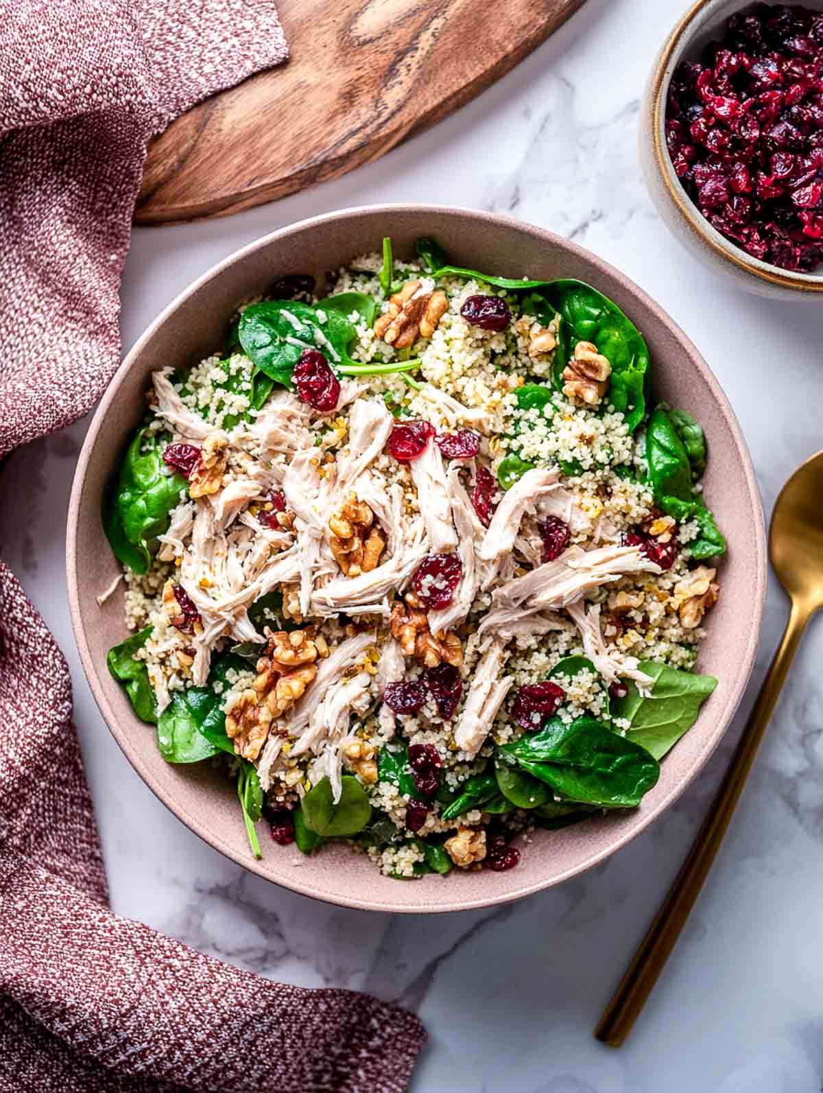 A top-down view of a winter chicken spinach salad topped with shredded chicken, fluffy cranberry orange couscous, toasted walnuts, and dried cranberries, served in a muted red bowl.