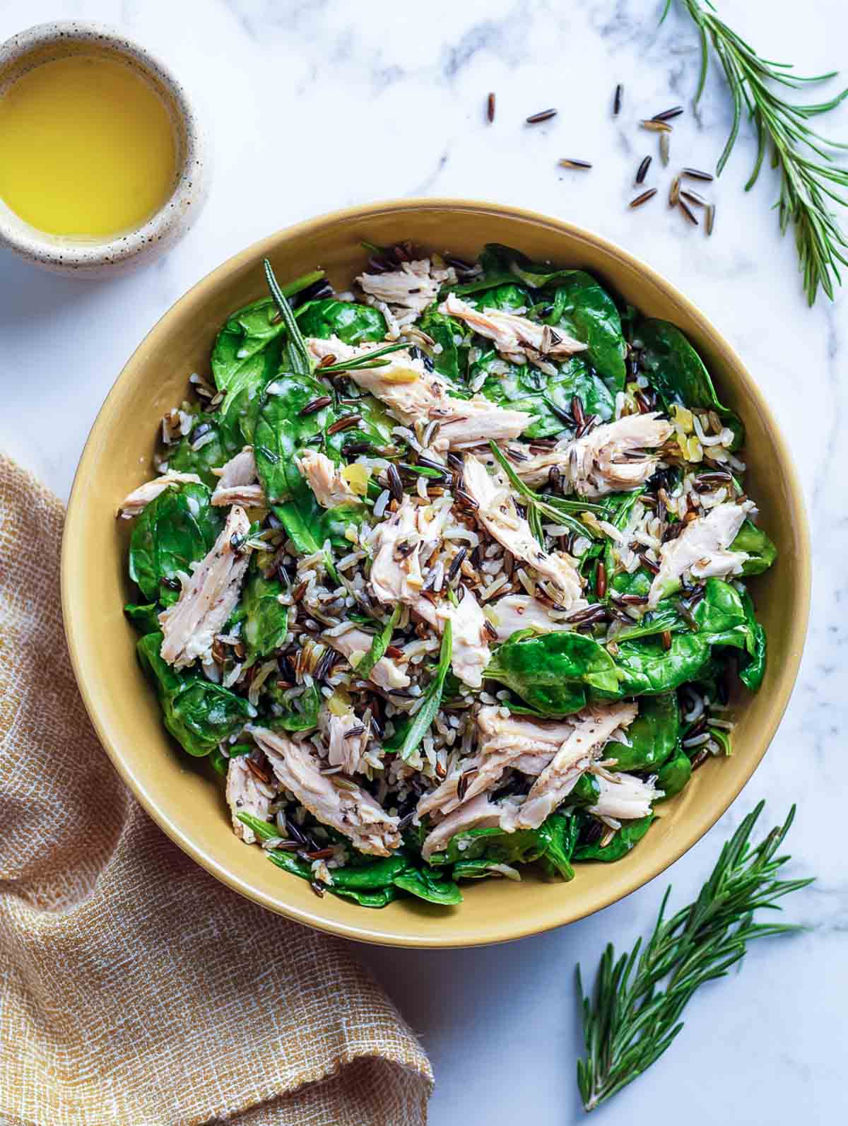A top-down view of a winter salad with shredded chicken, wild rice, fresh spinach, and rosemary arranged in a muted ceramic bowl on a marble surface.