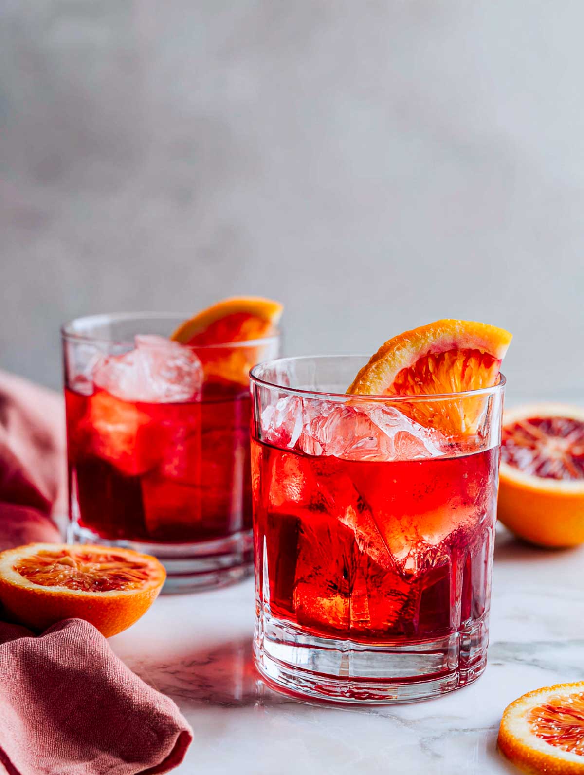 Side view of two blood orange negronis in rocks glasses on a white marble surface.
