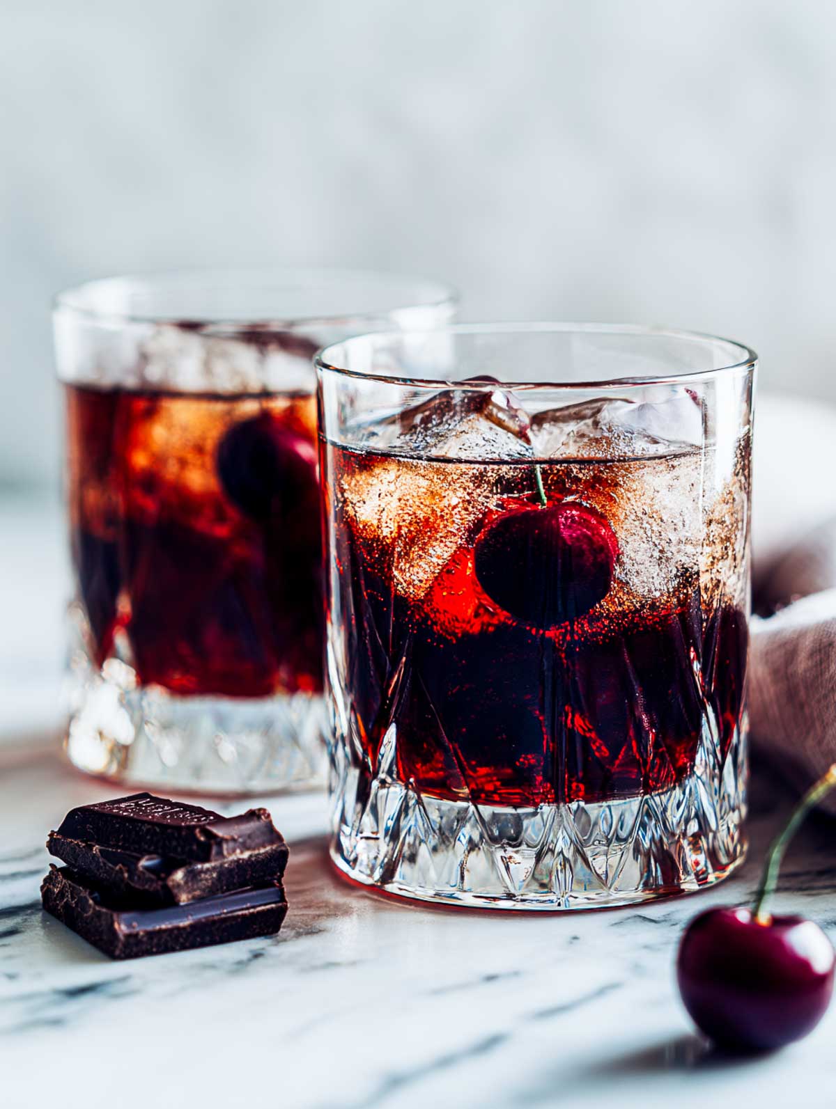 Side view of two dark chocolate cherry old fashioneds in rocks glasses on a white marble surface.