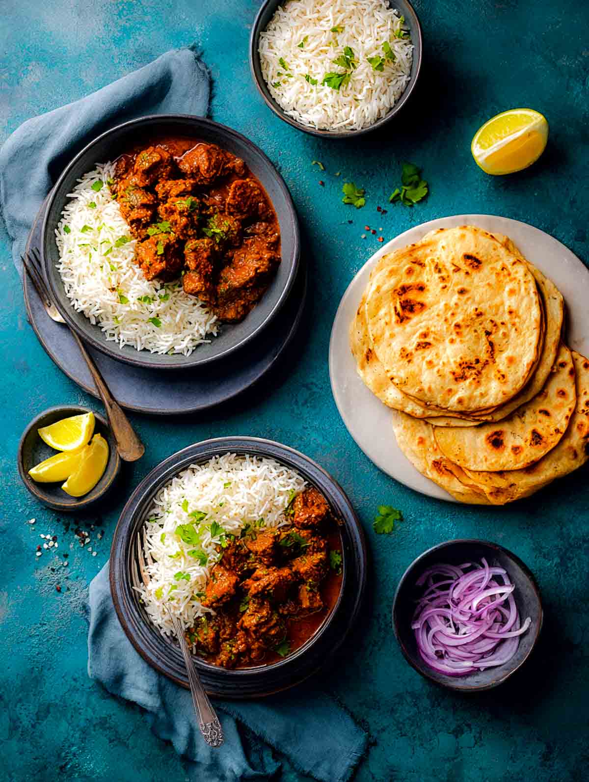 Two plated servings of lamb bhuna with basmati rice, roti, and simple side accompaniments.