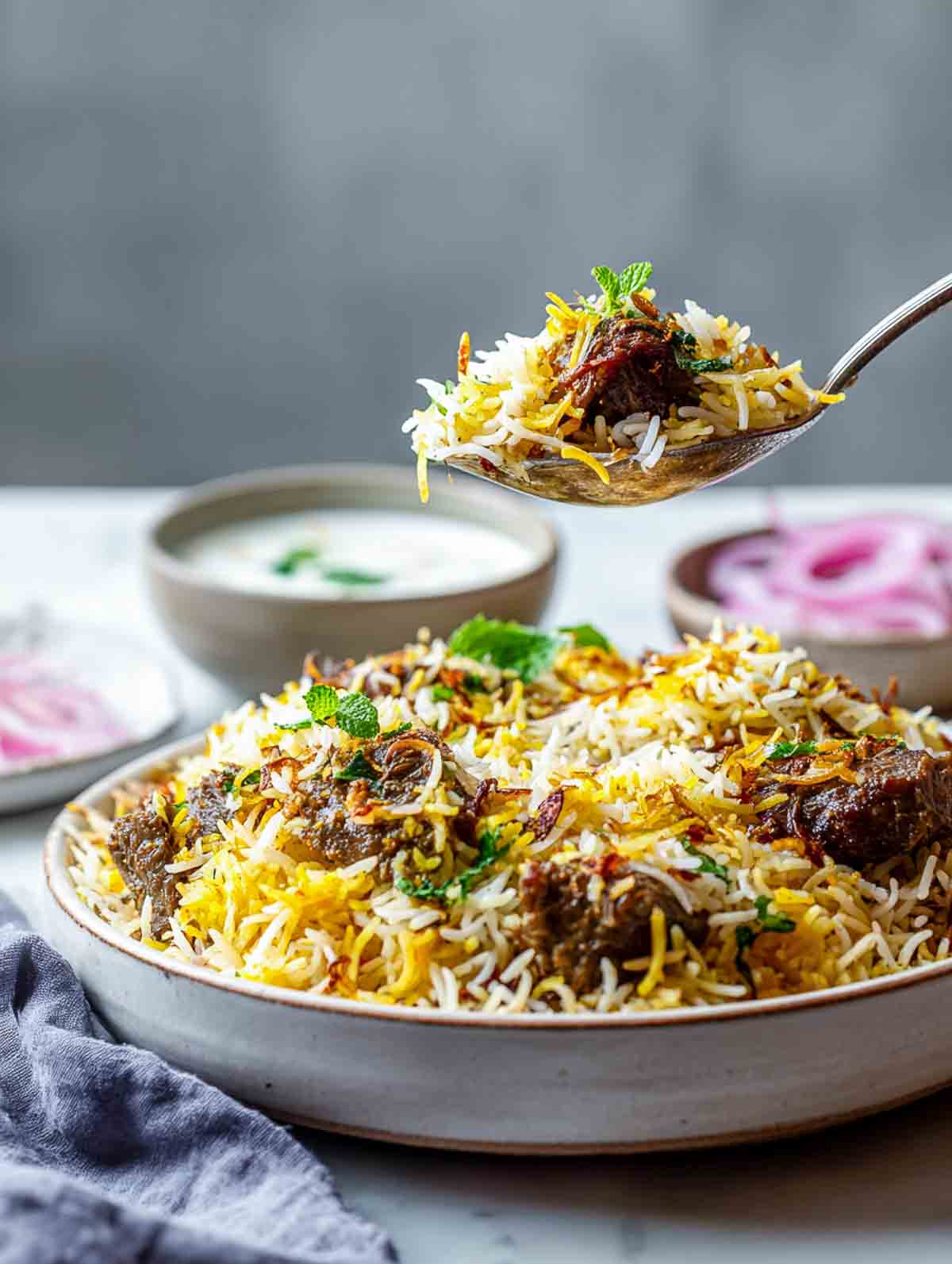 A spoon lifts tender lamb biryani from a serving bowl with raita.