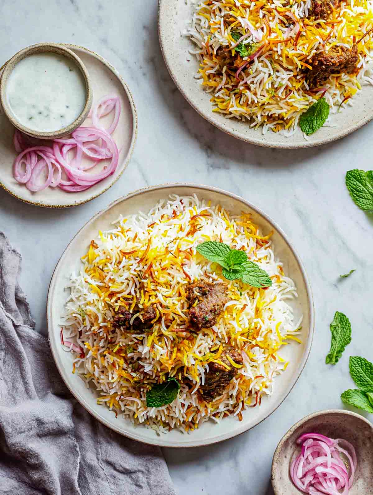 Two plates of lamb biryani arranged with raita and sliced onions.