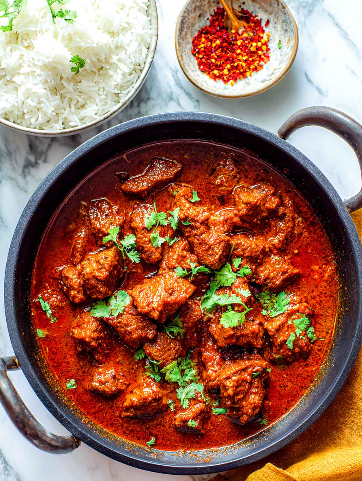 This image shows a black pan filled with Lamb Madras simmered in a deep red gravy, garnished with cilantro and served alongside fluffy basmati rice and chili flakes.