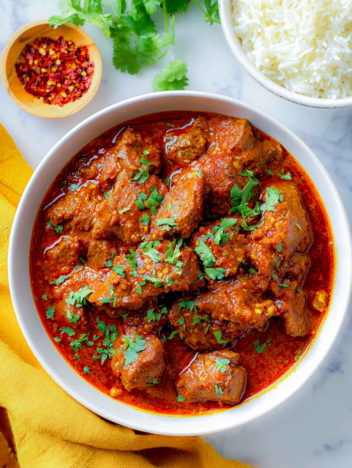 This photo features a bowl of Lamb Madras with tender lamb pieces coated in a rich red masala, garnished with cilantro and set on a marble background with rice and chili flakes nearby.
