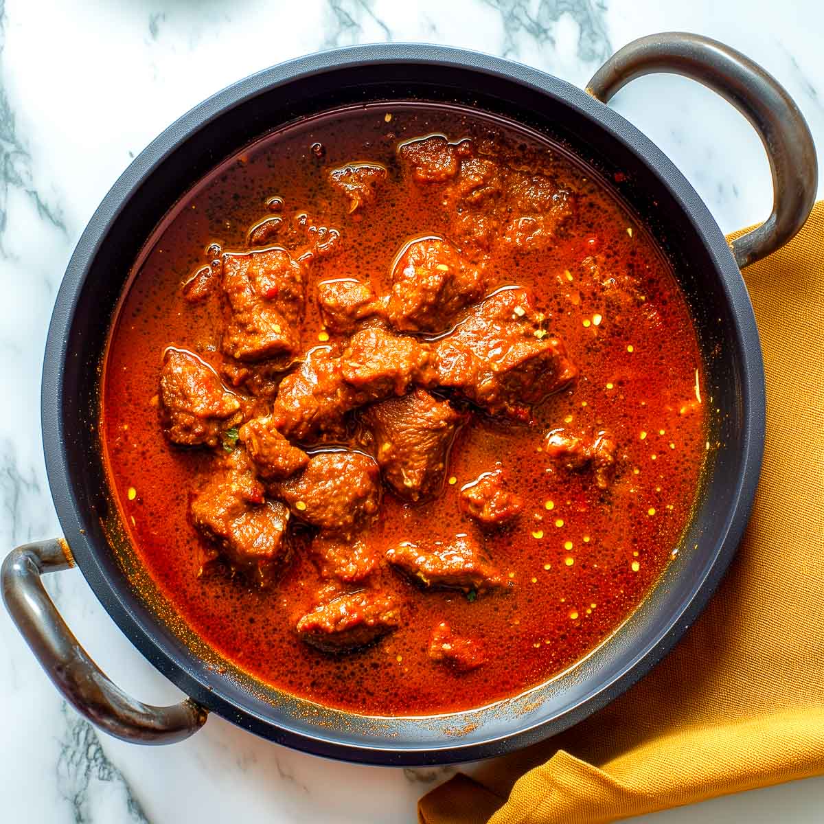 This photo shows Lamb Madras simmering in the pan with tender lamb pieces surrounded by a deep red-brown curry and flecks of oil on the surface.