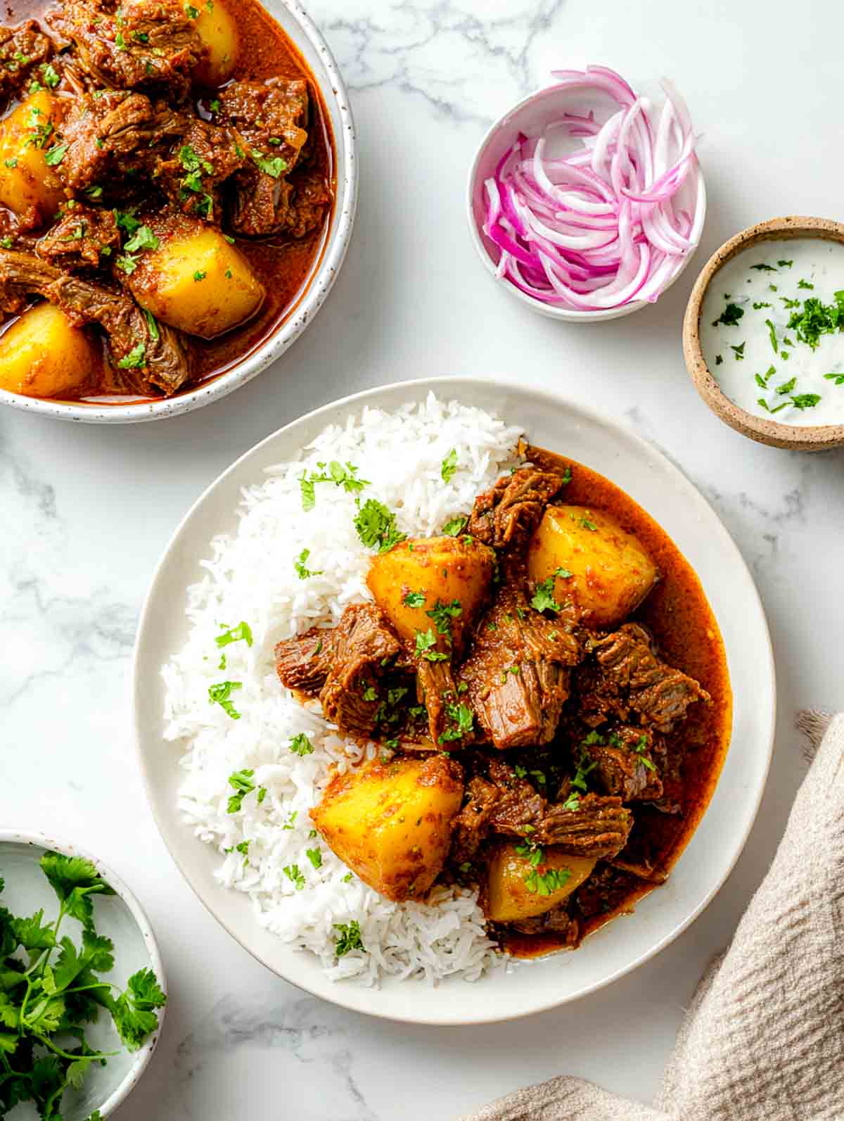 Plated lamb and potato curry with basmati rice, red onion slices, and yogurt.