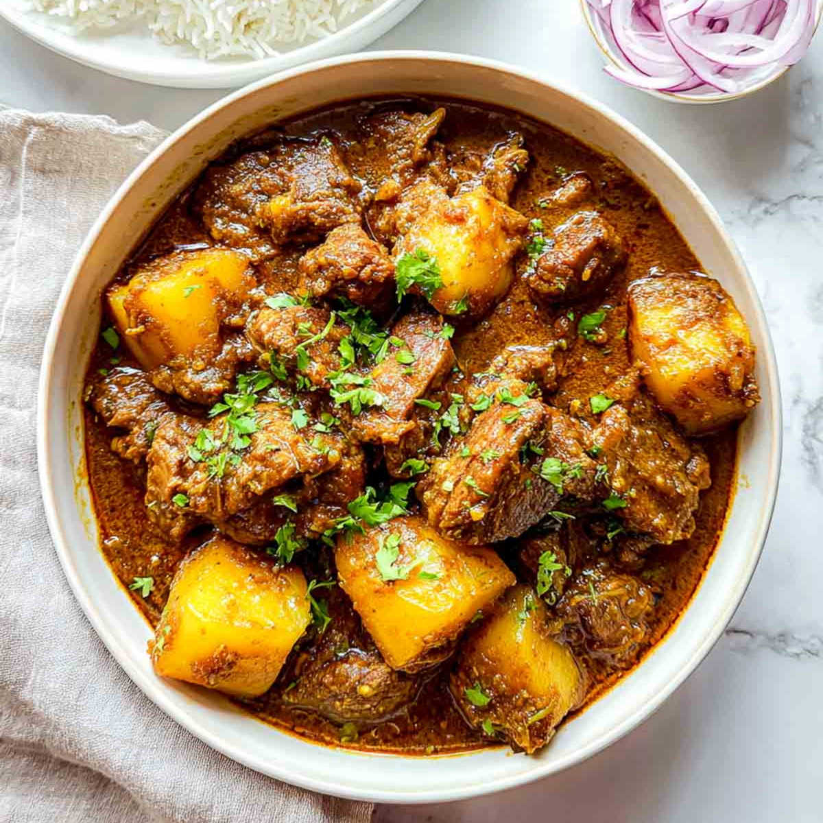 Closeup view of lamb and potato curry with in a serving bowl with rice and sliced onions in the background.