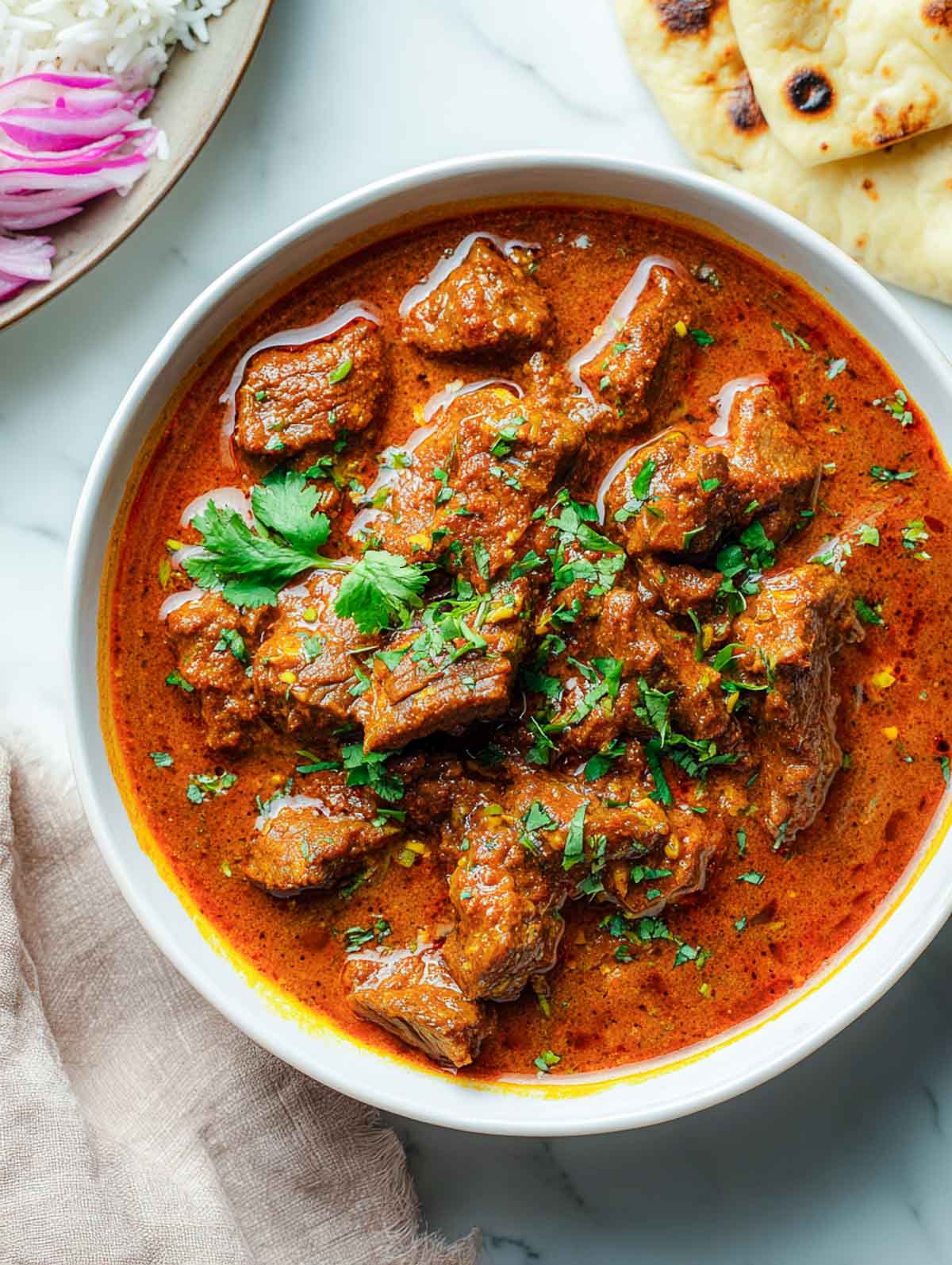 A serving bowl filled with lamb rogan josh is garnished with fresh cilantro, with rice and naan visible in the background.