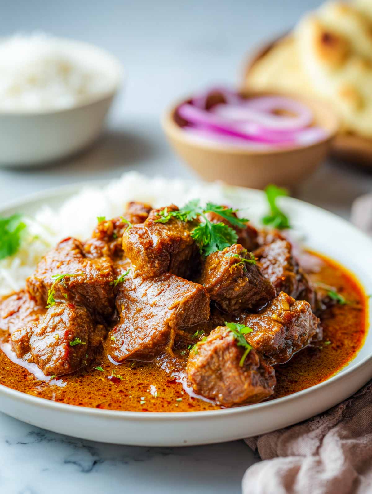 A plated serving of lamb rogan josh is shown over rice, with sliced onions and naan blurred in the background.