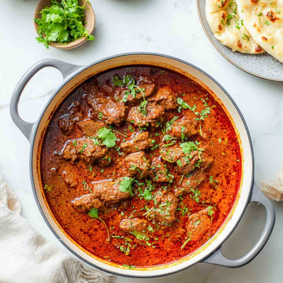 The lamb rogan josh is presented in a Dutch oven with a rich red gravy and fresh cilantro sprinkled on top.