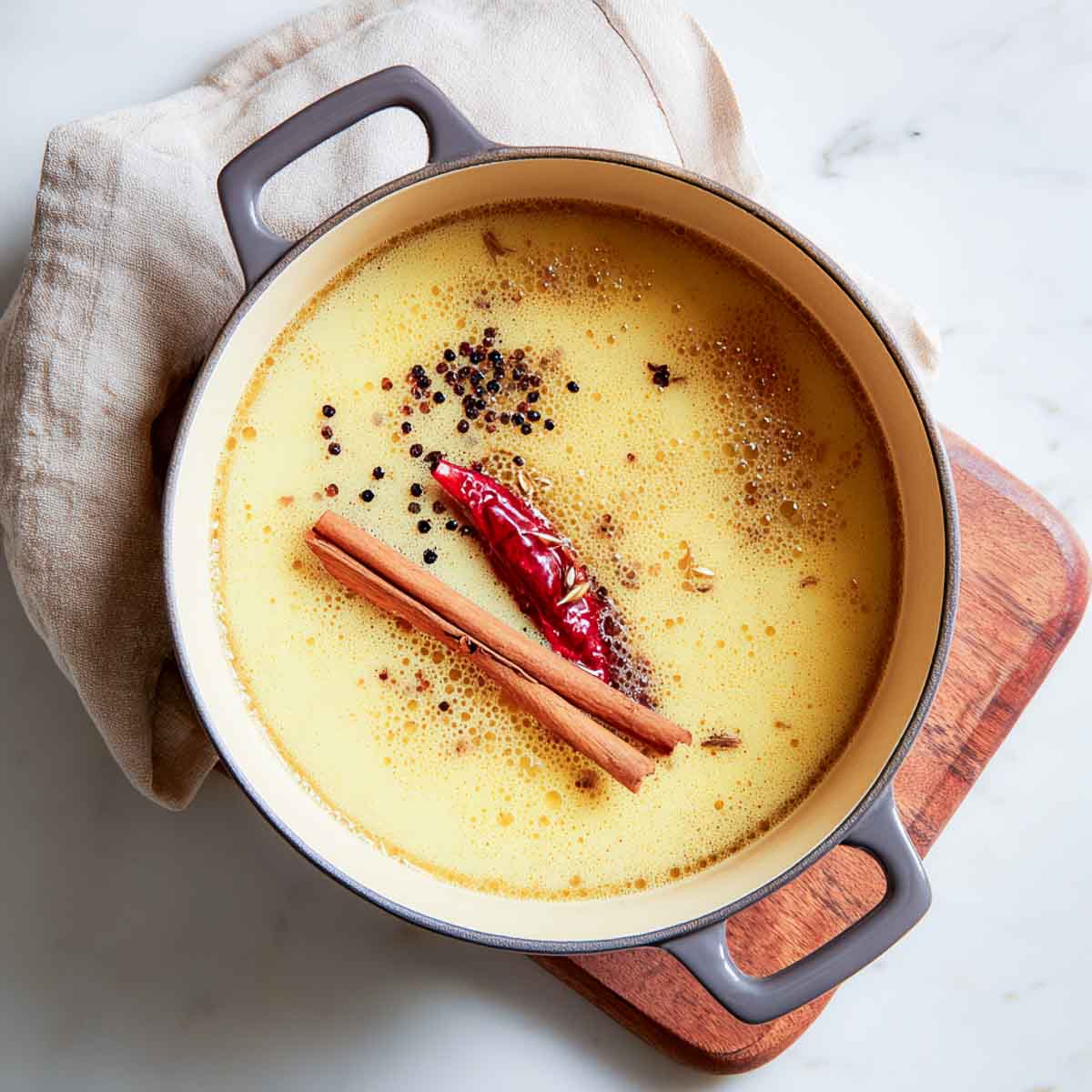Whole spices including cinnamon, dried chili, peppercorns, and fennel are simmering in a Dutch oven filled with foamy spiced ghee.