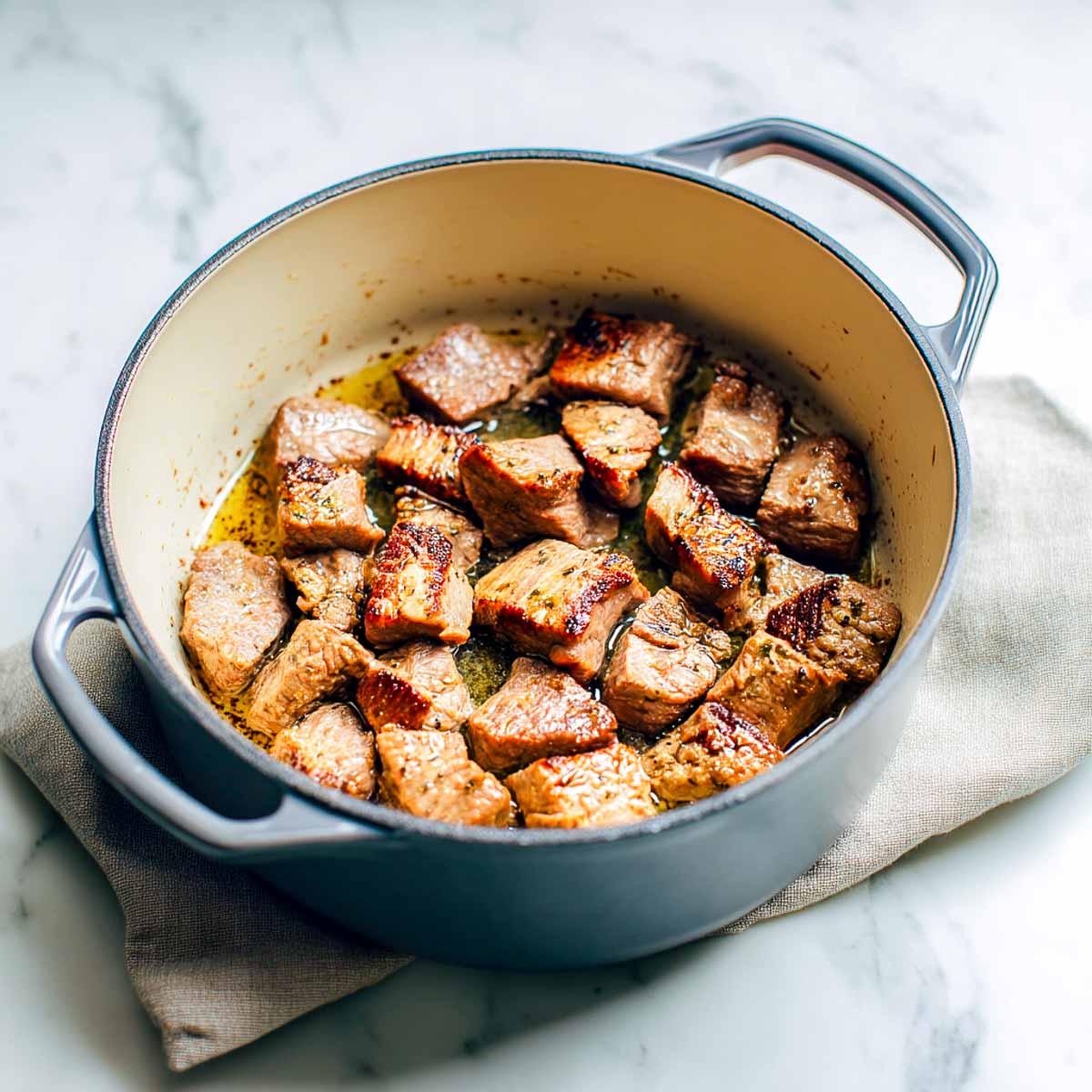 Lamb pieces are browning in a grey Dutch oven, developing a golden sear on all sides.