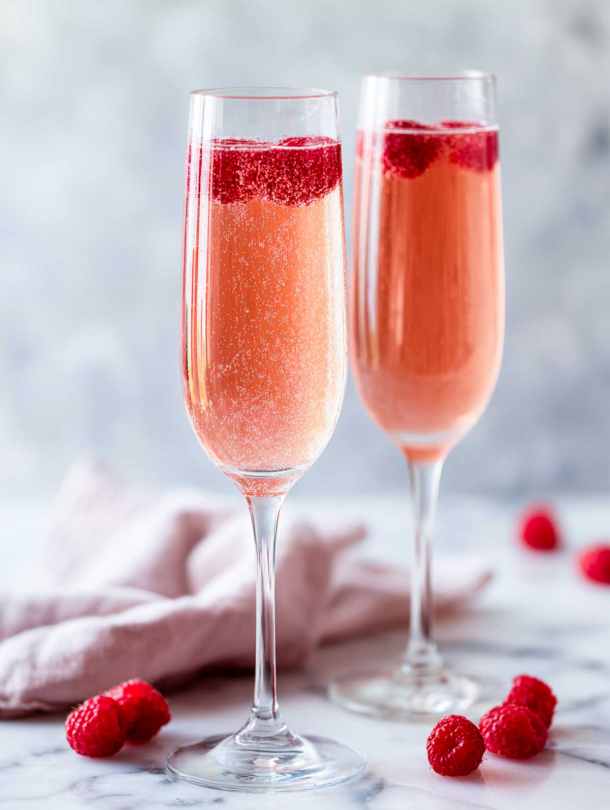 Side view of two raspberry French 75 cocktails in champagne flutes on a white marble surface.