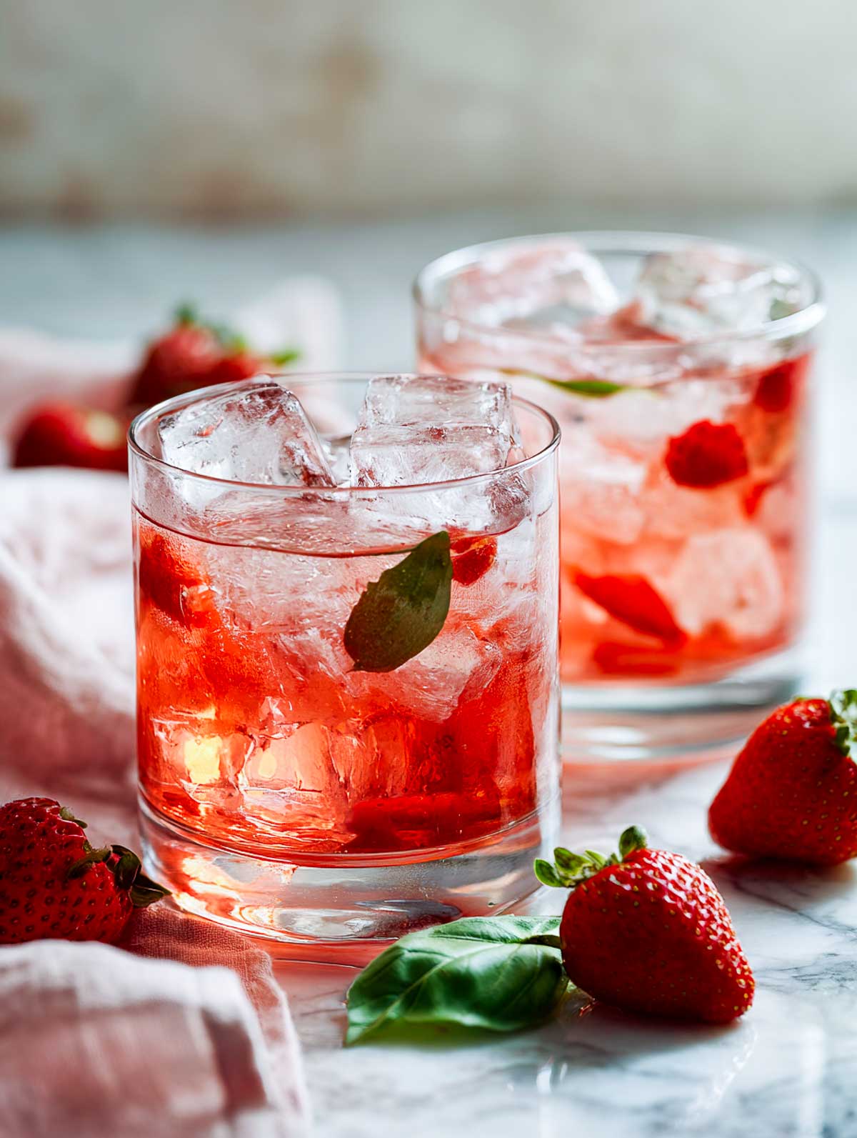 Side view of two strawberry basil rum smash cocktails in rocks glasses on a white marble surface.