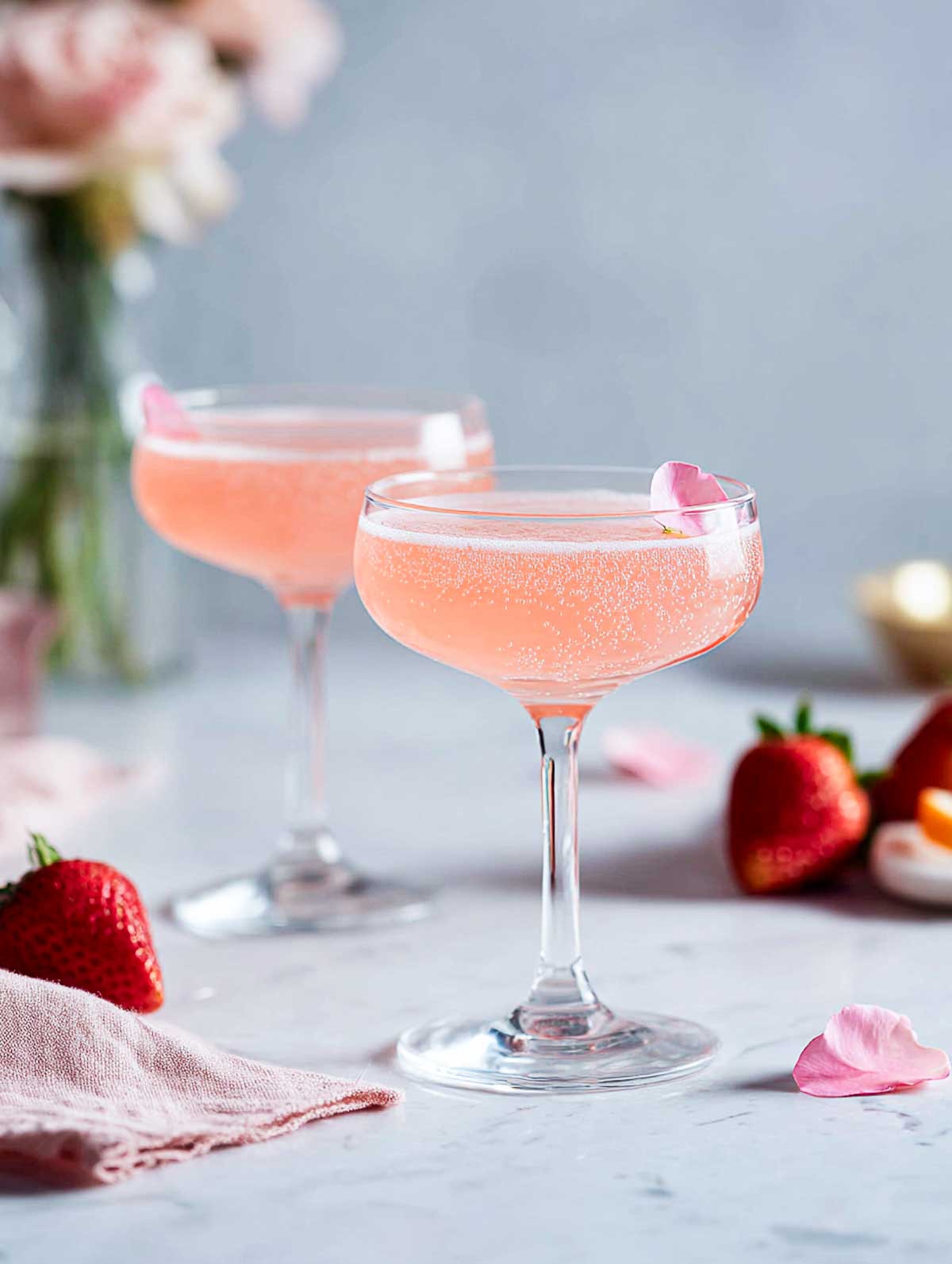 Side view of two strawberry rose gin fizzes in coupe glasses on a white marble surface.