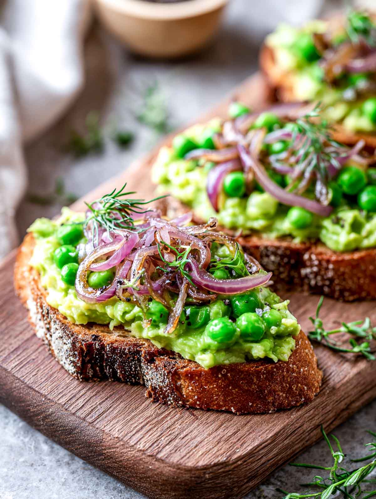Slices of rustic toast topped with creamy avocado and pea mash and finished with pickled shallots and fresh herbs on a wooden board.